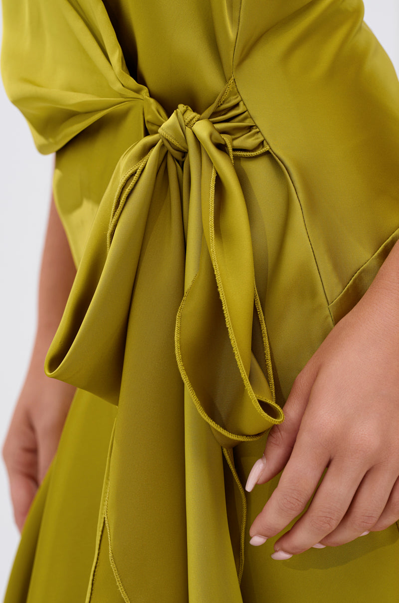 A hand rests near a large, eye-catching bow at the waist of the STAR OF THE SHOW SATIN MINI DRESS, featuring smooth, flowing olive-green satin fabric.
