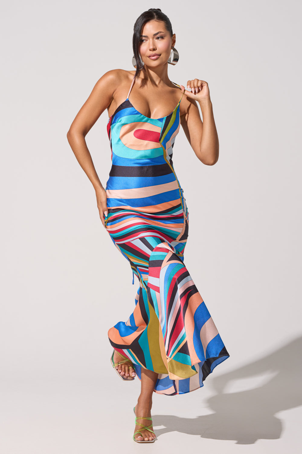 Model wears the 13 GOING ON 30 MAXI DRESS, featuring a colorful abstract print with bold geometric patterns and a flared hem. She pairs it with green heeled sandals, large hoop earrings, and side-swept hair.
