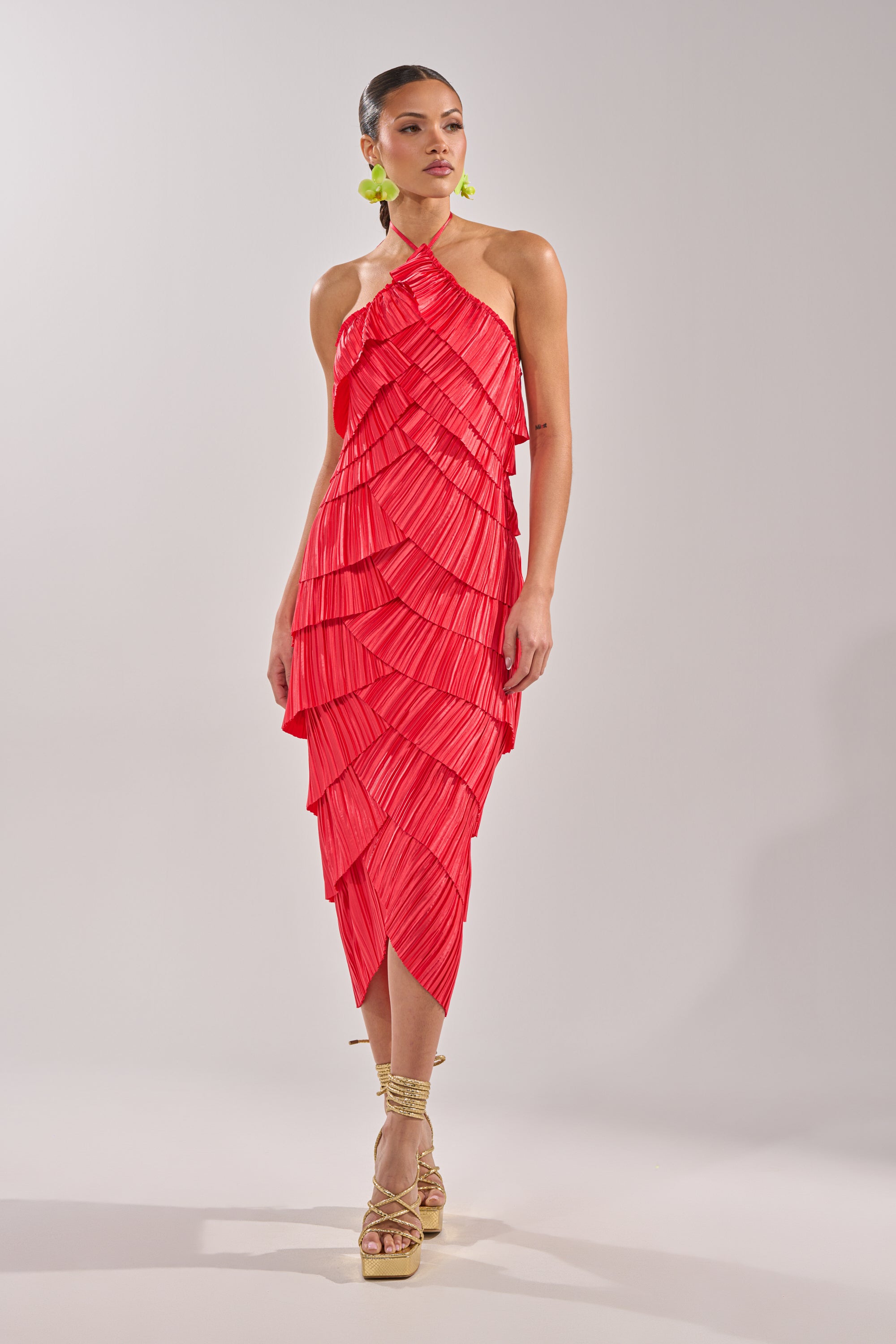 A woman wears the GIVE IT TO ME BABY SATIN MAXI DRESS—a bright red, halter-neck, layered silhouette with accordion pleats and scalloped satin. Her hair’s pulled back, complemented by green earrings and strappy beige platform sandals. 1