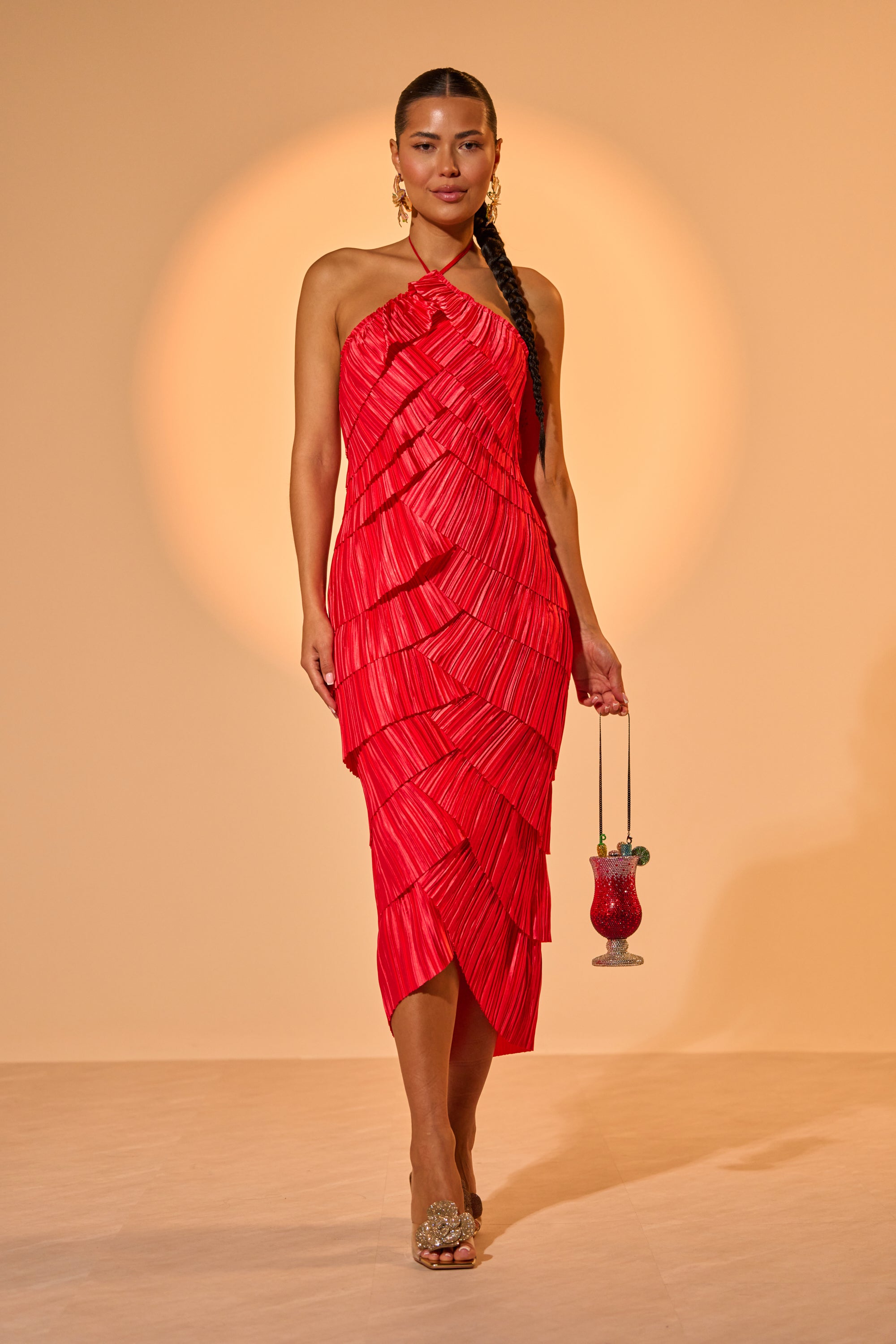 A woman stands confidently in the GIVE IT TO ME BABY SATIN MAXI DRESS, a vibrant red pleated satin maxi, paired with clear heels and a small decorative bag against a warm, softly lit background. 0