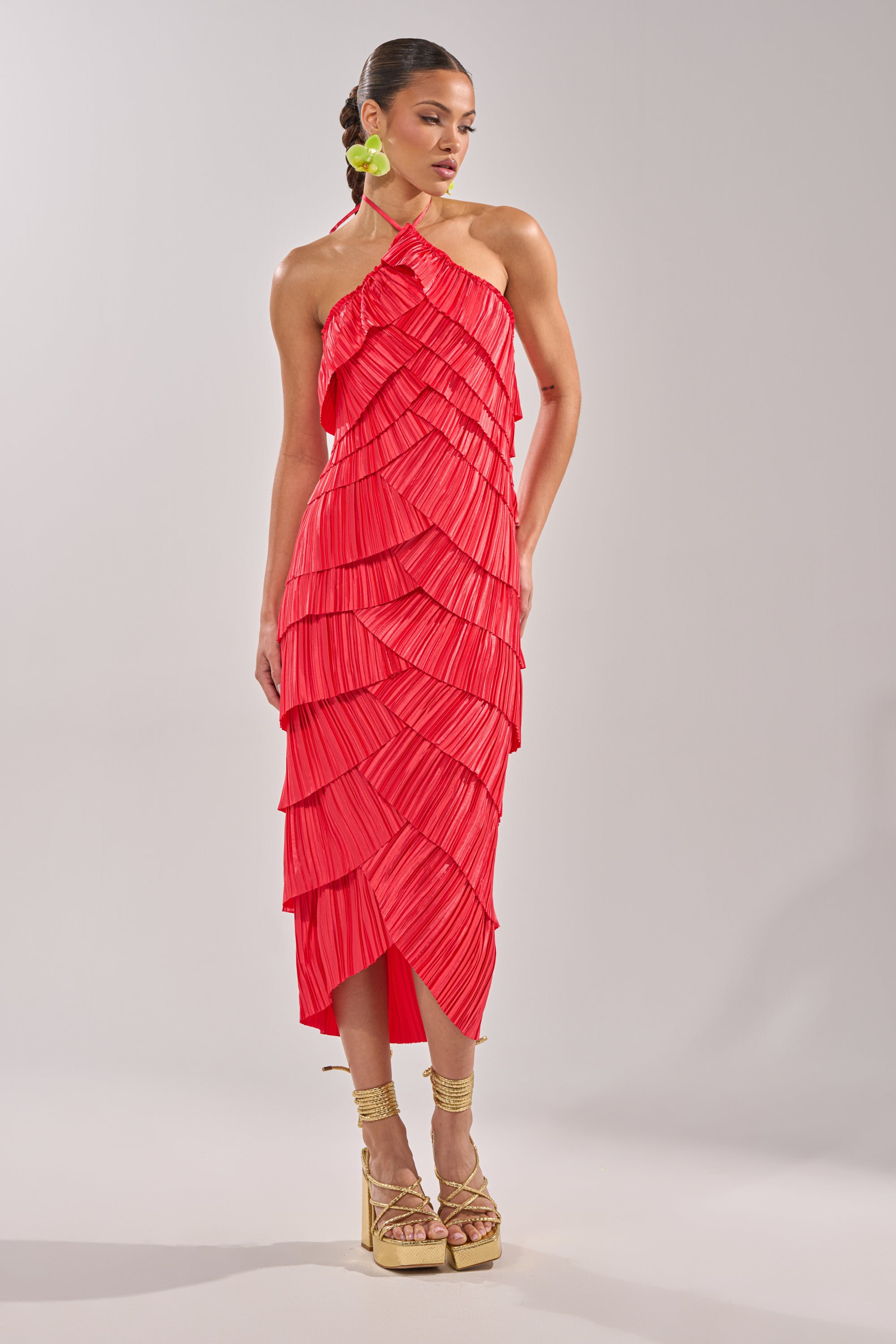 A woman stands in a neutral studio wearing the GIVE IT TO ME BABY SATIN MAXI DRESS, a sleeveless, one-shoulder bright red satin dress with accordion pleats and layered ruffles, paired with gold high platform sandals and green round earrings. 2