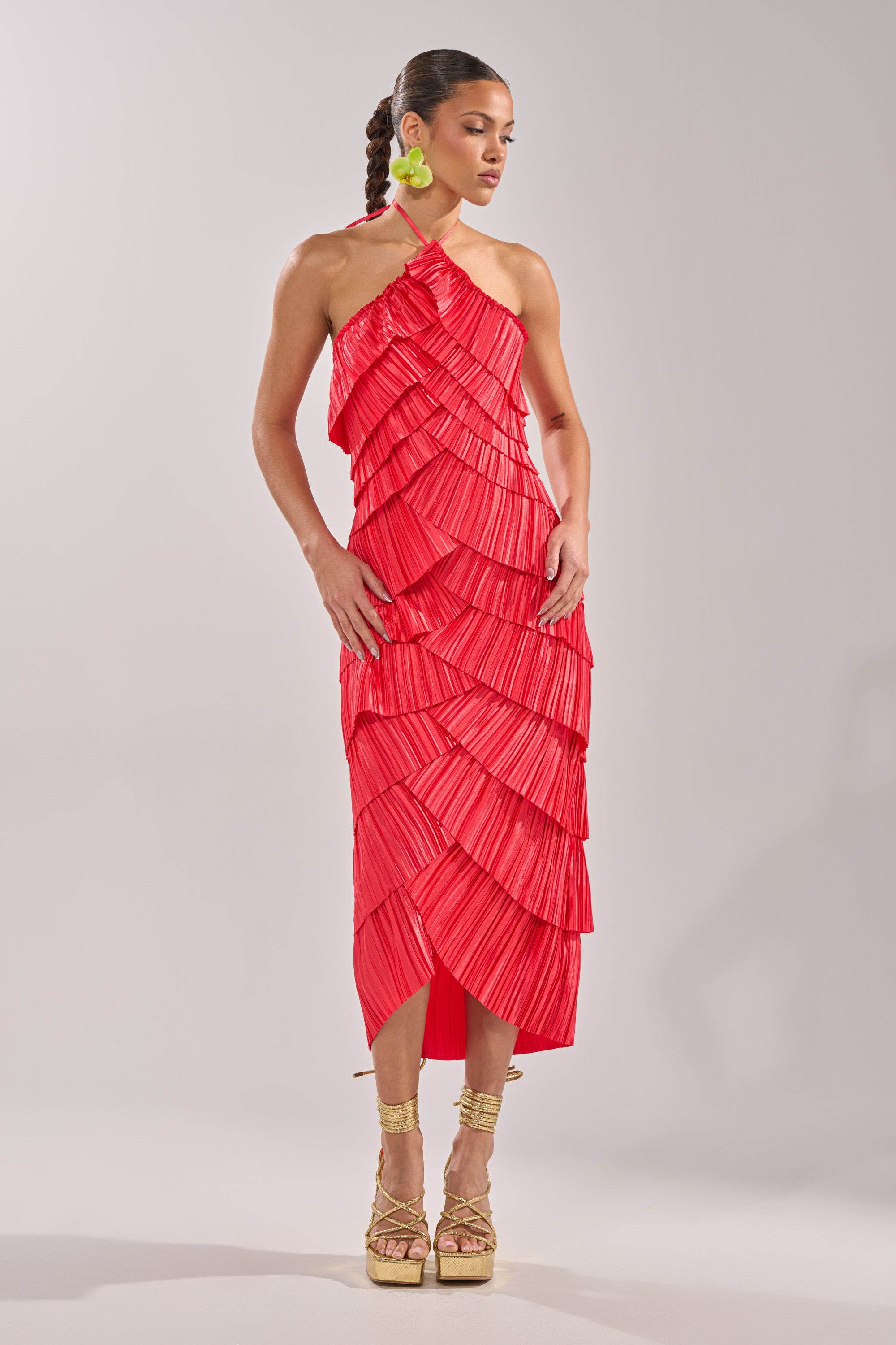 A woman poses in the GIVE IT TO ME BABY SATIN MAXI DRESS, a bright red pleated maxi with a halter neckline and asymmetrical hem. She wears braided hair, green statement earrings, and gold platform sandals against a plain light background. 4