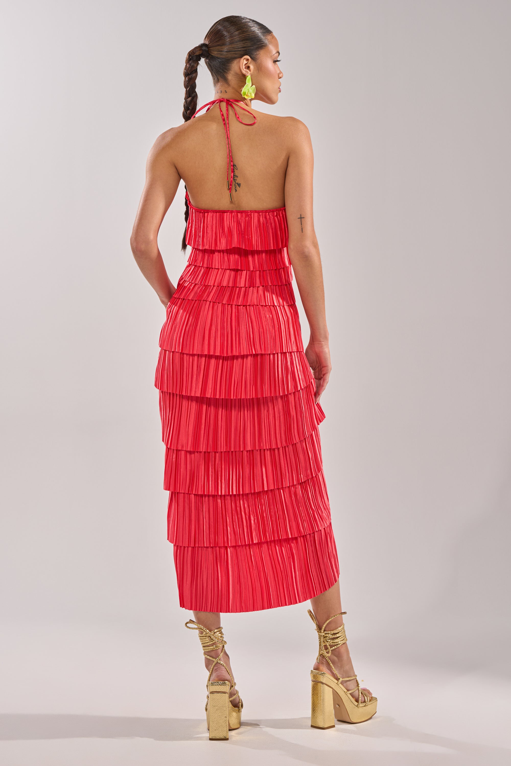 A woman faces away, wearing the GIVE IT TO ME BABY SATIN MAXI DRESS—a pleated red halter-neck maxi with open back. Her long braid, gold earrings, and beige platform sandals with ankle straps complete the look against a light background. 6
