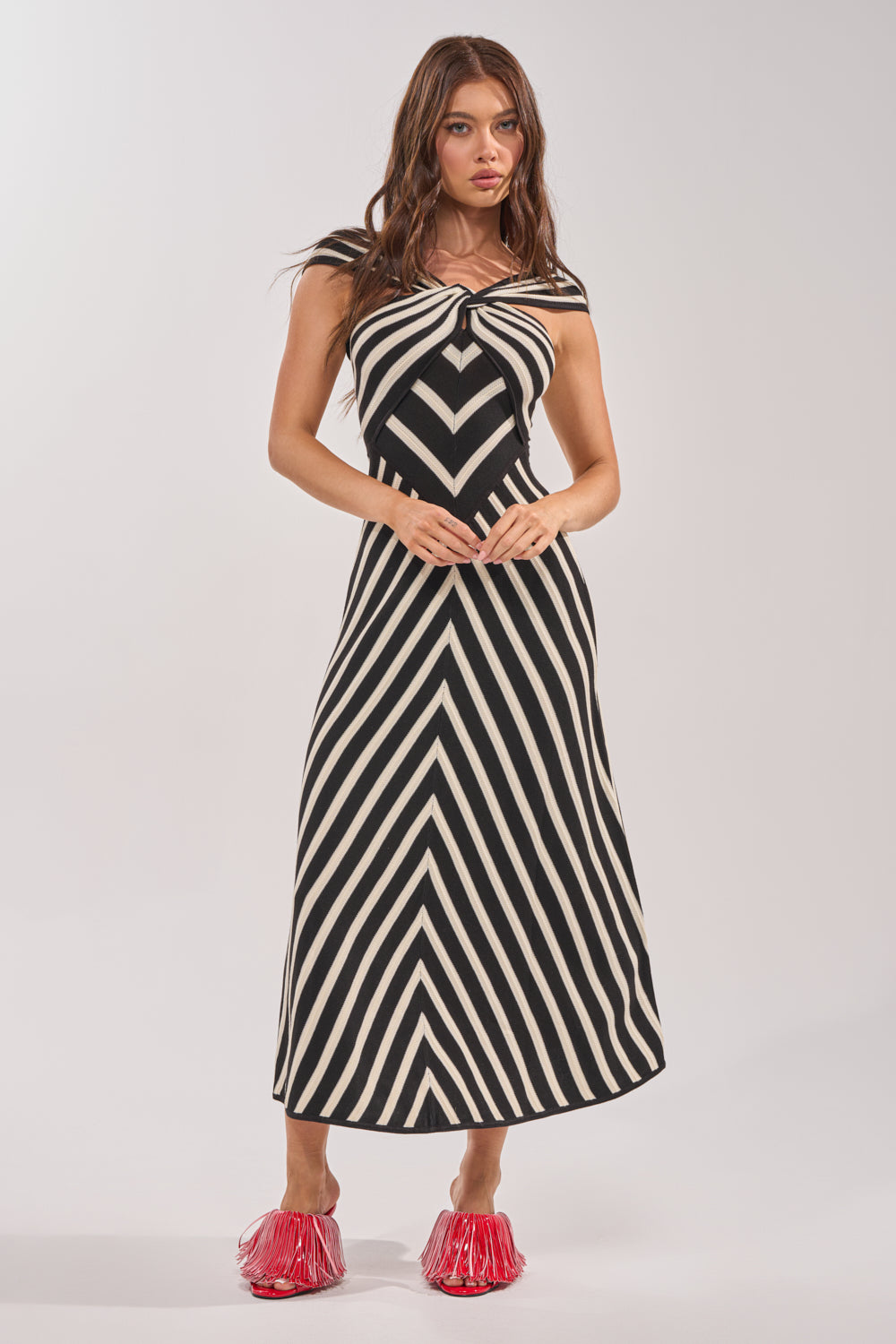 A woman wears the BLOCKED AND BLESS MAXI SWEATER DRESS IN BLACK AND WHITE STRIPE, a sleeveless maxi with bold chevron patterns, paired with bright red slip-on shoes against a plain light background. 2
