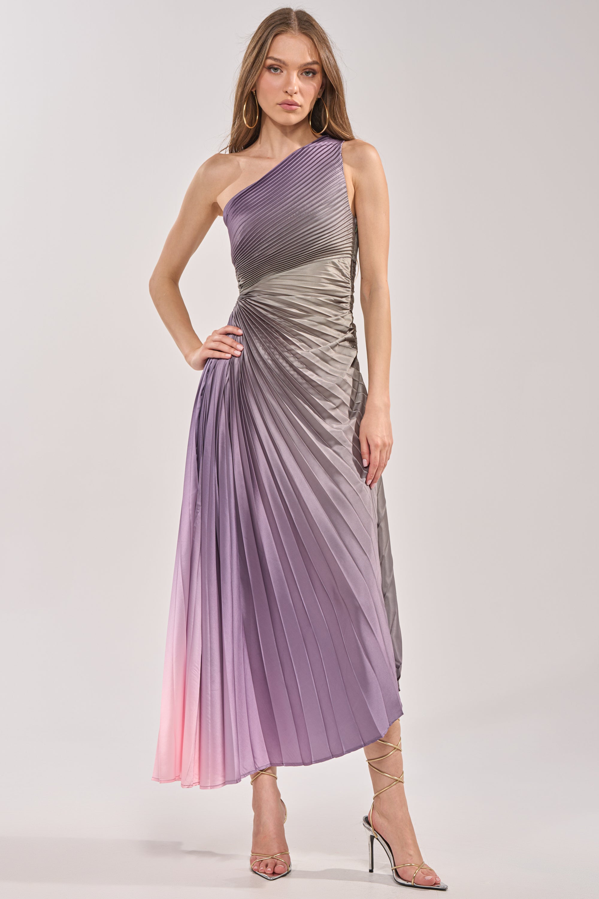 A woman stands in the HERE TO SHINE PURPLE MAXI DRESS, a satin pleated ombré one-shoulder maxi fading from gray to light purple and pink. She pairs it with strappy high-heeled sandals, her long hair flowing loose. 1