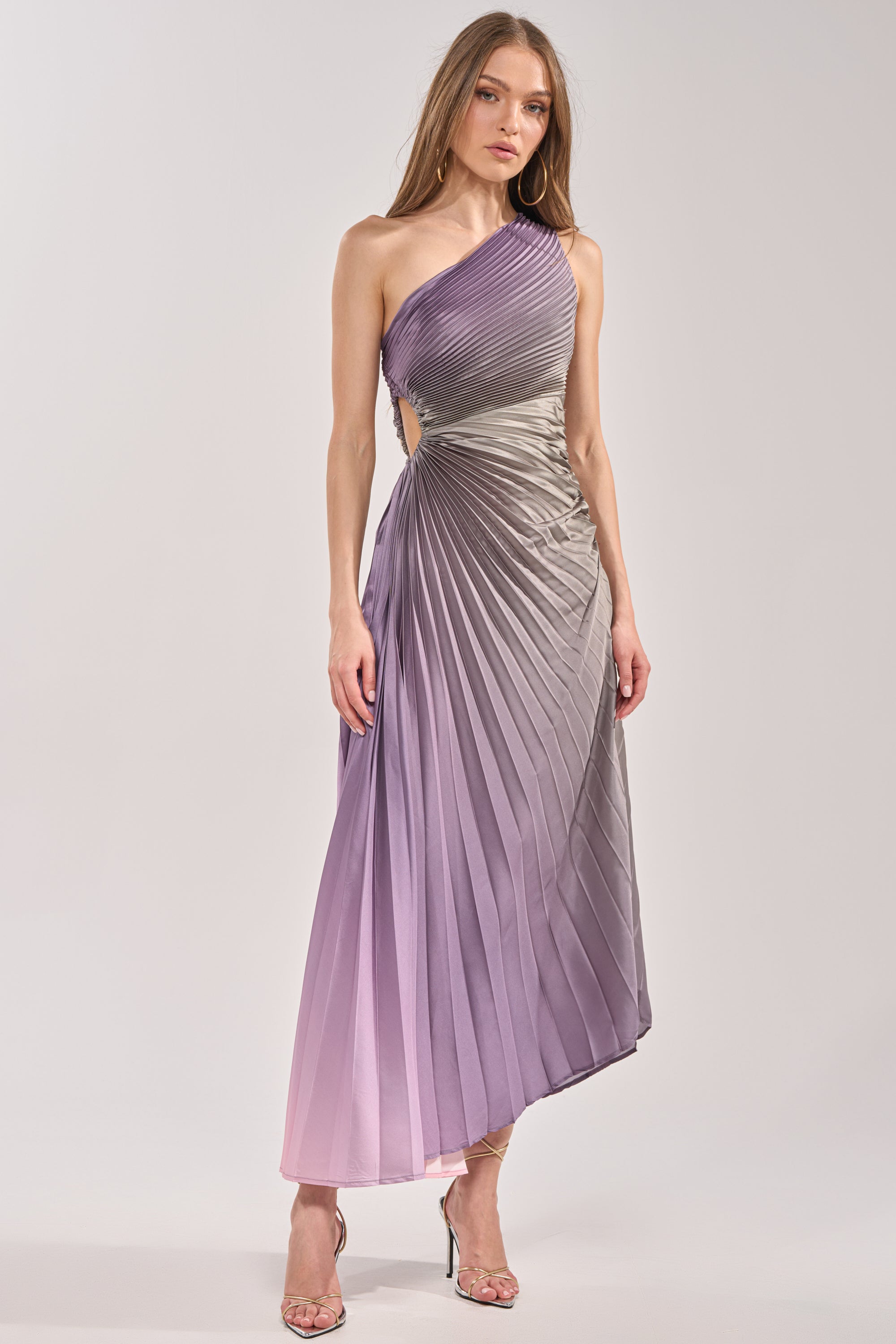 A woman stands against a plain background wearing the HERE TO SHINE PURPLE MAXI DRESS— a satin pleated maxi in gradient purple and gray, styled with hoop earrings, long straight hair, and strappy high-heeled sandals. 2