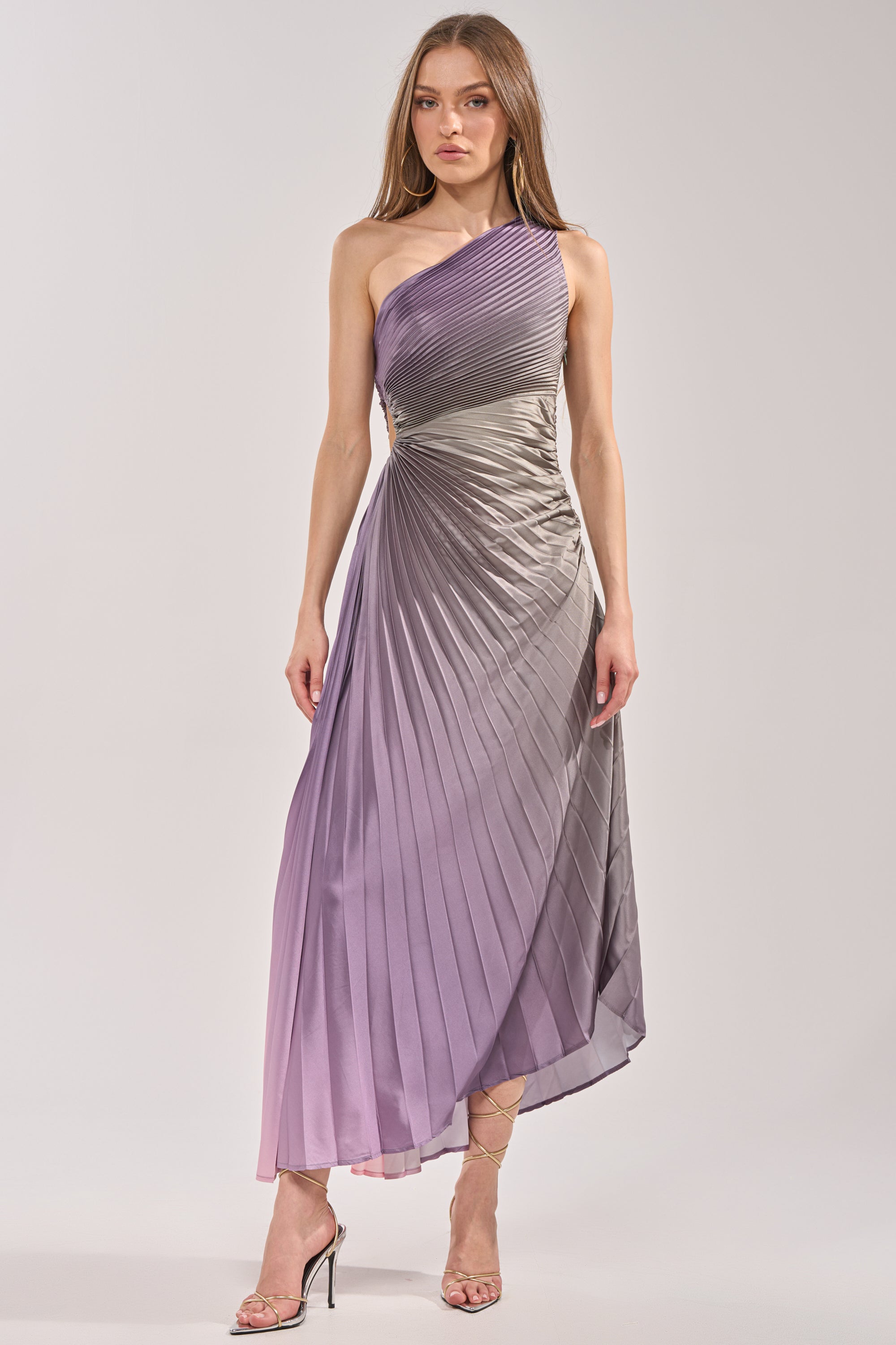 A woman in the HERE TO SHINE PURPLE MAXI DRESS, a sleeveless satin pleated maxi with gradient gray to lavender hues, pairs it with high-heeled sandals and stands against a plain light background. 3