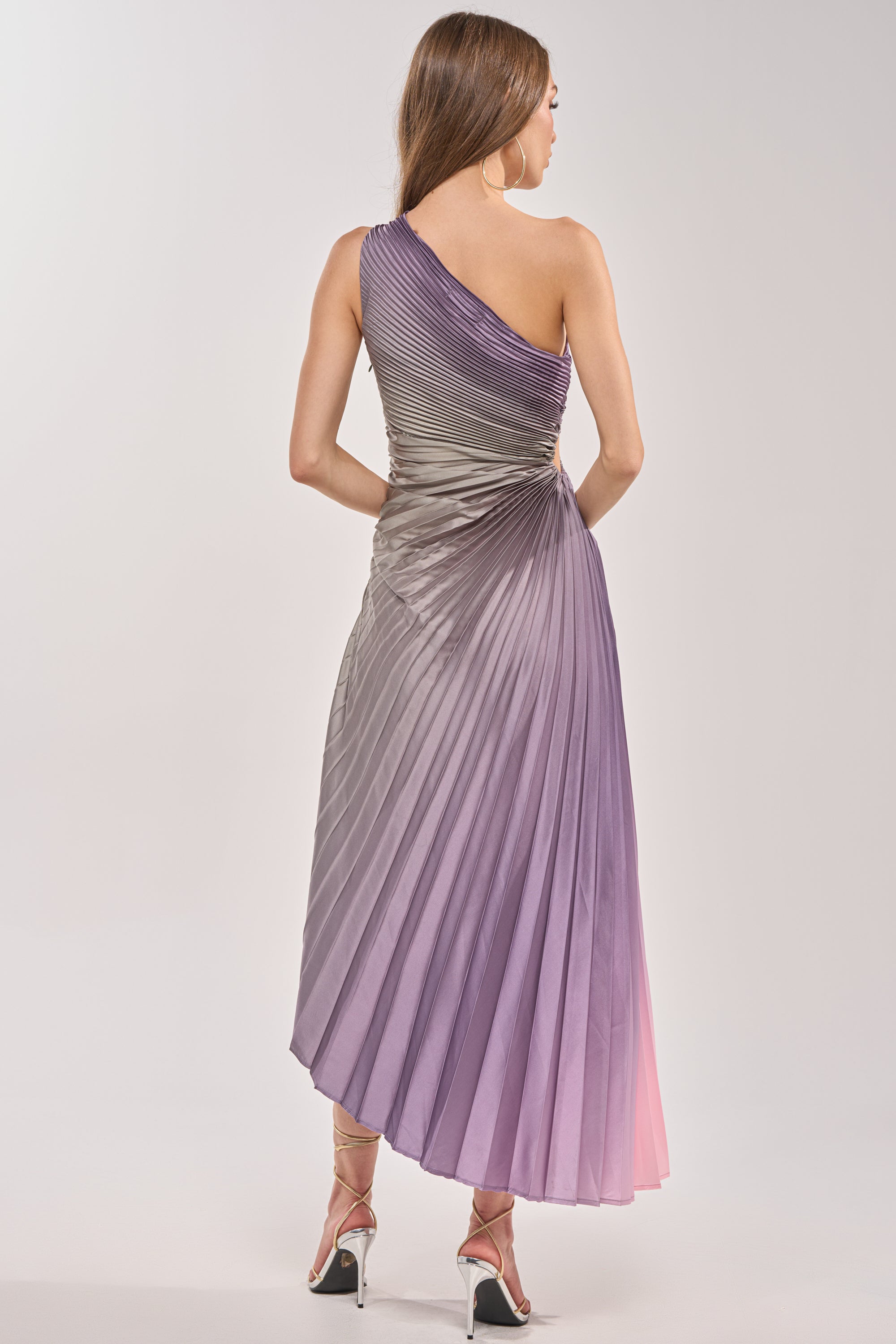 A woman, seen from behind, wears the HERE TO SHINE PURPLE MAXI DRESS—a one-shoulder ombré gown in pleated purple and pink shades—paired with high-heeled sandals. 5