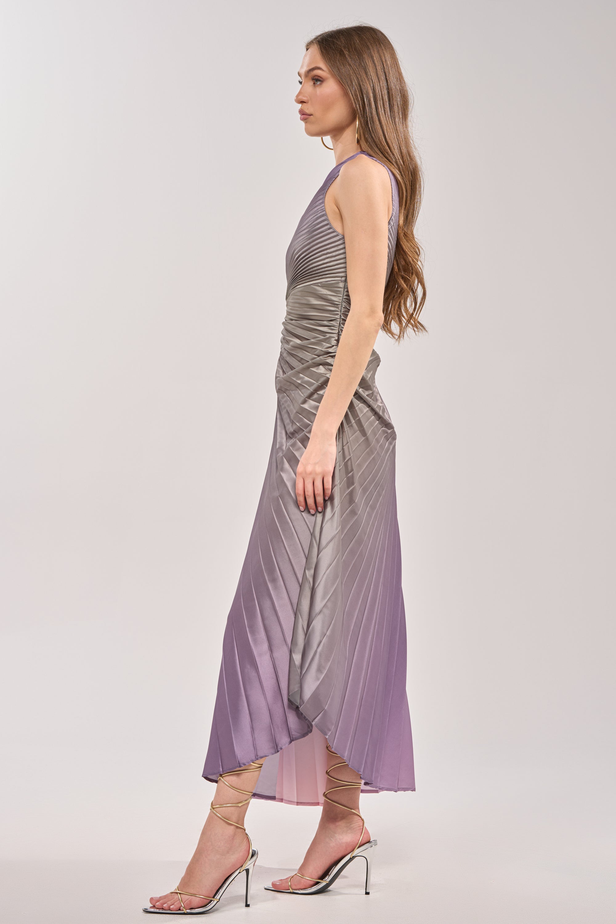 A woman poses in profile in the HERE TO SHINE PURPLE MAXI DRESS, featuring gray and lavender ombré pleats, styled with strappy heels. Her long, wavy hair is worn loose against a simple backdrop. 6