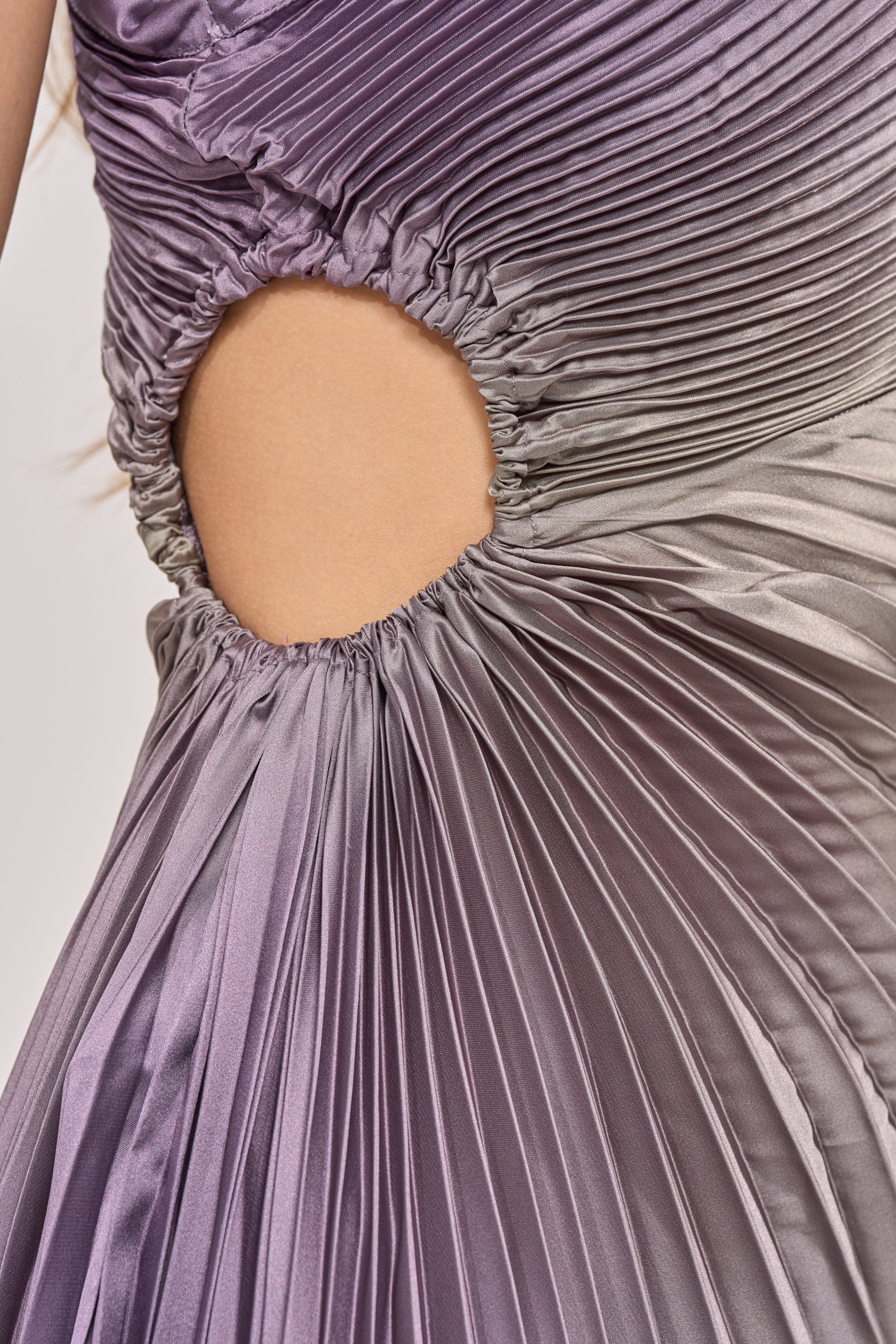 Close-up of the HERE TO SHINE PURPLE MAXI DRESS, a formal ombré gown in iridescent purple and gray with pleated satin fabric, round waist cutout, and gathered ruching that enhances its silhouette for an elegant evening look. 8