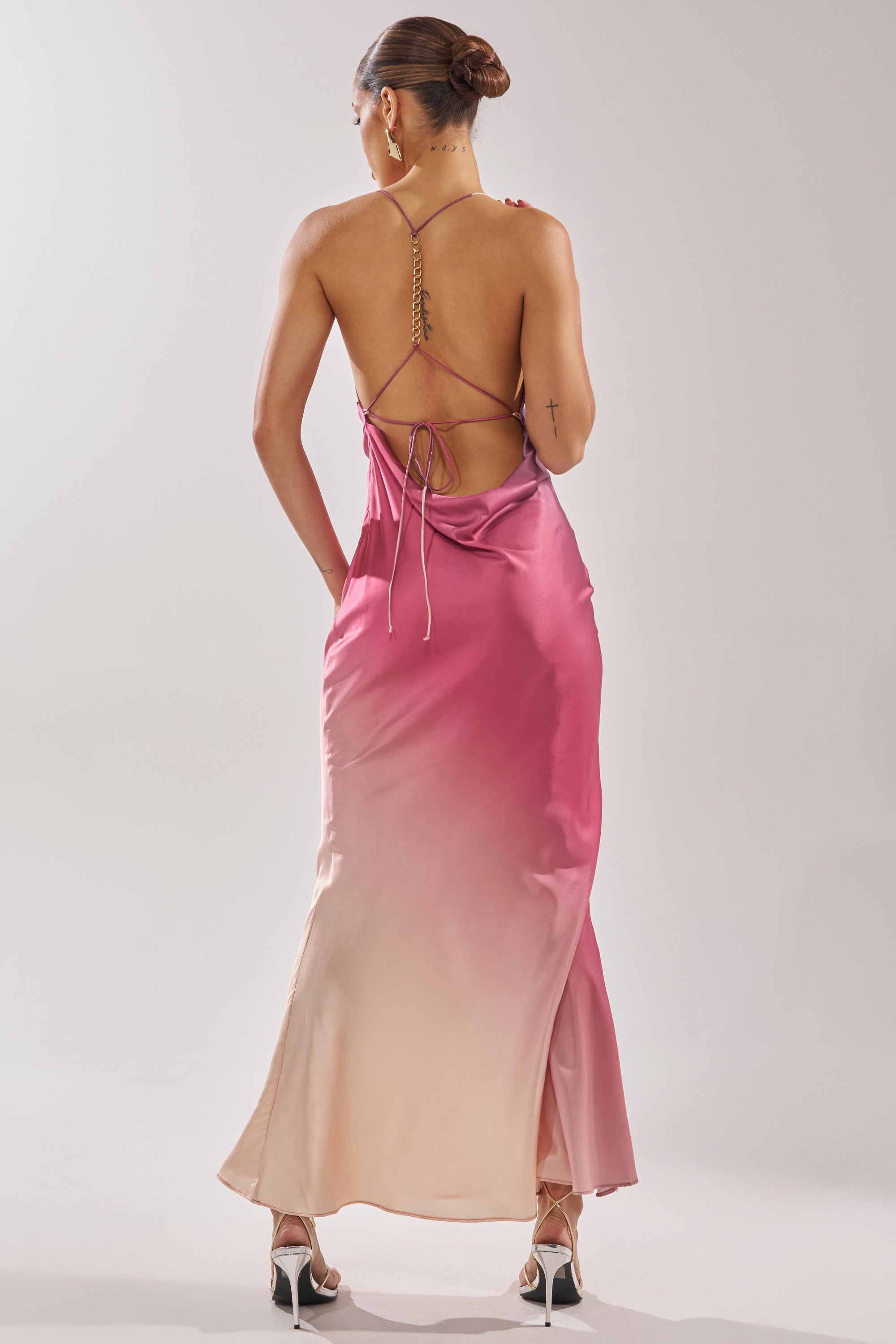 A woman with her hair in a bun wears the PRETTY WOMAN MAUVE MAXI DRESS with thin crisscross straps and white high-heeled sandals, standing against a plain light background. 4