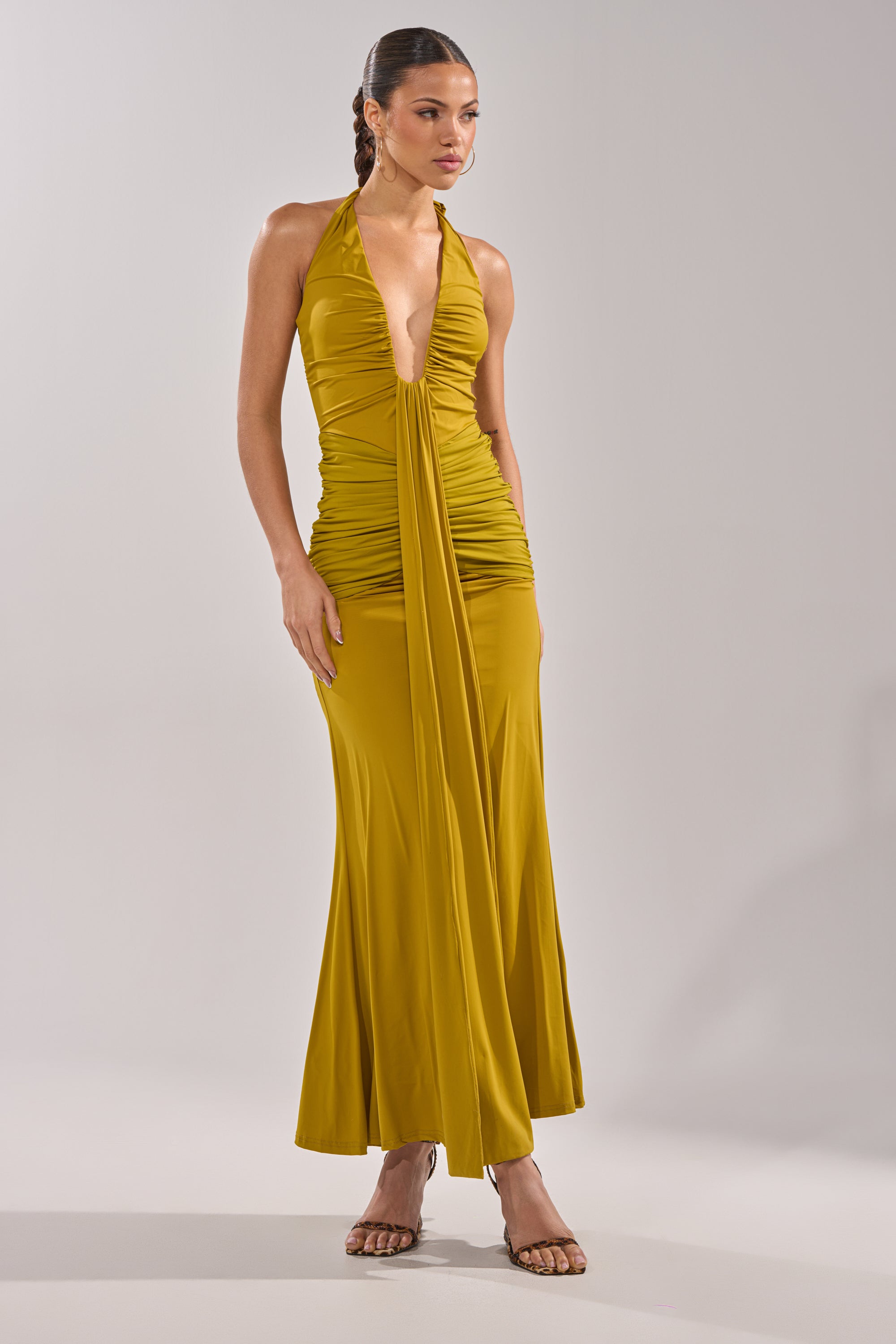 A woman wears the ISLAND LOVE MAXI DRESS IN GREEN, a halter-style maxi with a deep V-neckline, ruched bodice, draped sash, and flared floor-length hem, styled with strappy high-heeled sandals. 3