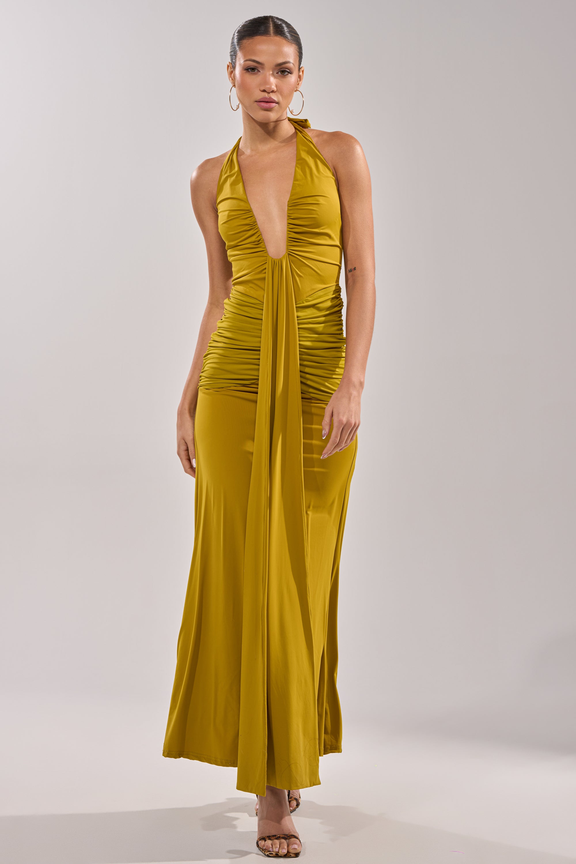 A woman wears the ISLAND LOVE MAXI DRESS IN GREEN, a halter-style maxi with a deep plunging neckline, ruched waist detailing, and a flowing floor-length skirt, standing against a plain light background. 0