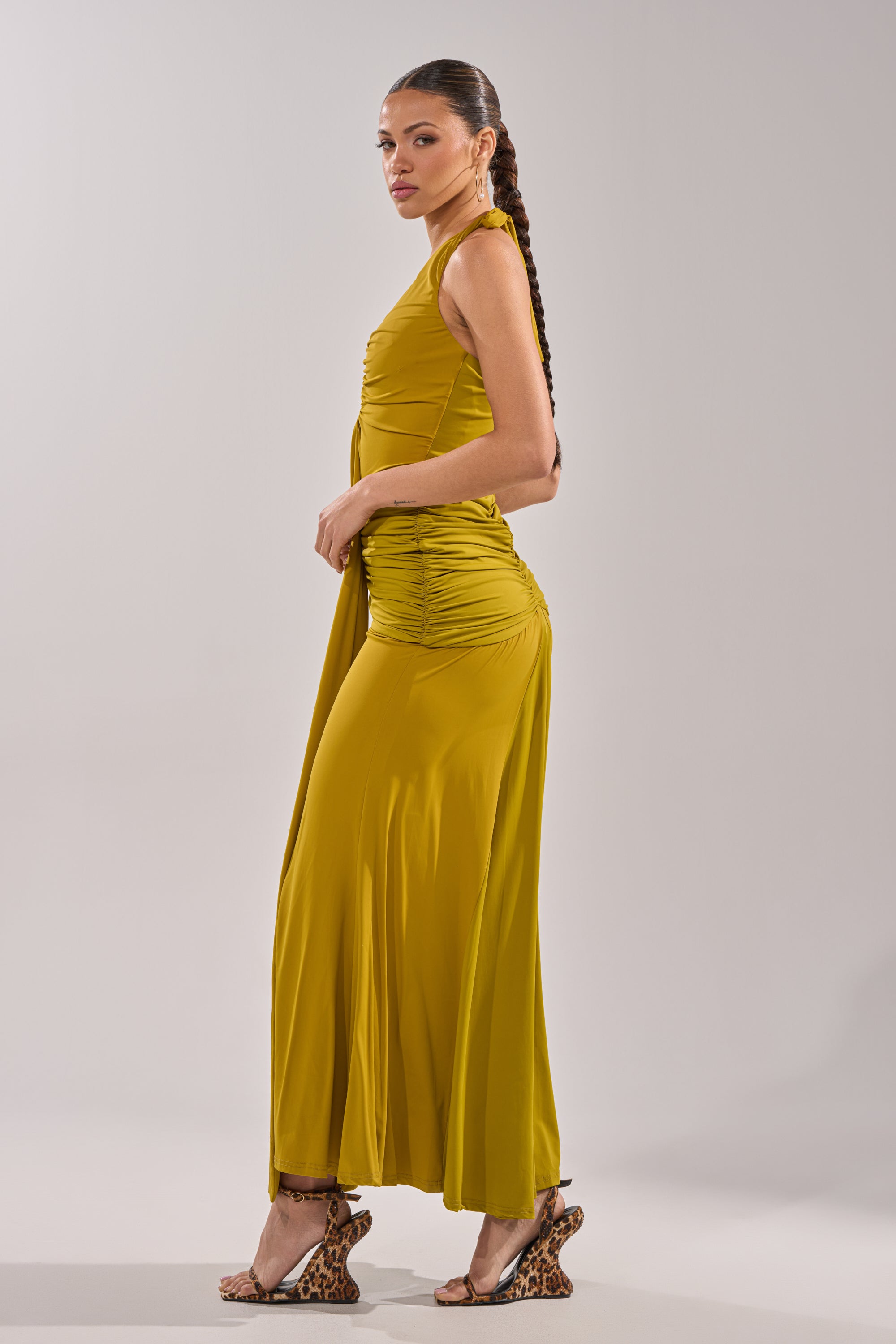 A woman with a long braid wears the ISLAND LOVE MAXI DRESS IN GREEN and leopard-print heeled sandals, standing sideways and looking over her shoulder against a plain light background. 6