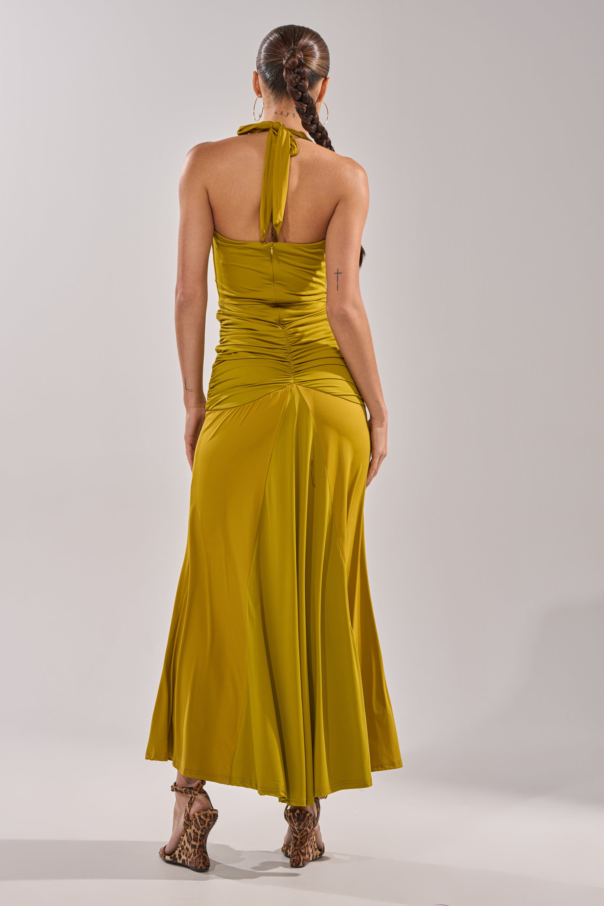 A person with a long braided ponytail stands with their back to the camera against a plain background, wearing the ISLAND LOVE MAXI DRESS IN GREEN and leopard print high-heeled sandals. 7