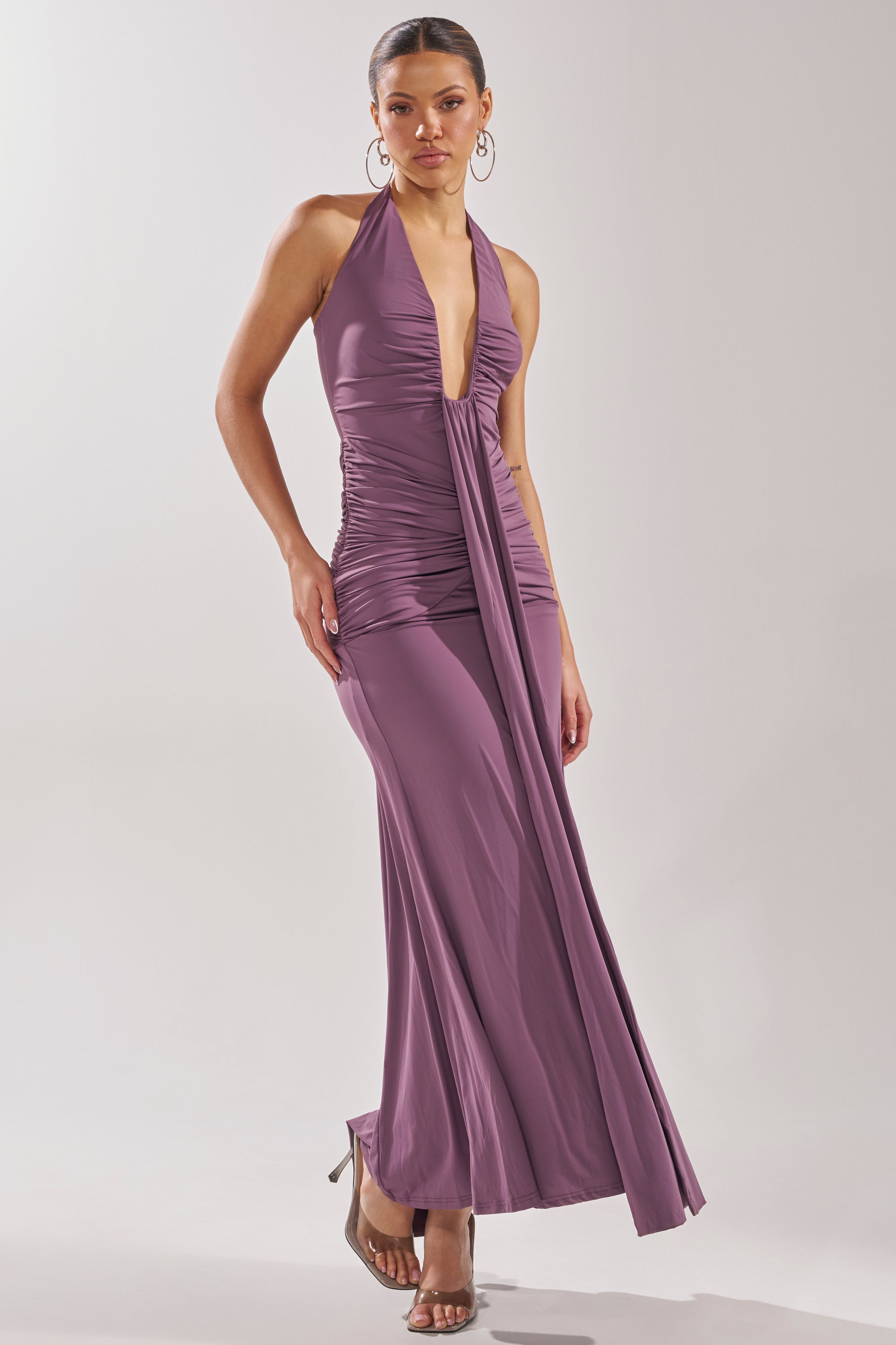 A woman wears the ISLAND LOVE MAXI DRESS in mauve, featuring a sleeveless halter neck, deep V-neckline, ruched bodice, and flowing skirt. She stands in high-heeled sandals against a plain light background—the perfect vacation dress. 1