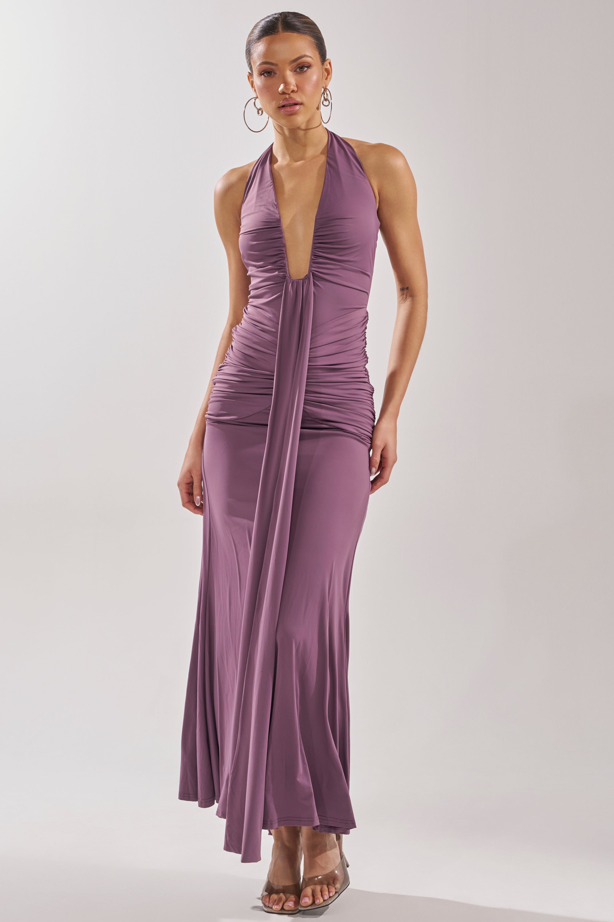A woman wears the ISLAND LOVE MAXI DRESS, a vacation-ready mauve halter maxi with a plunging neckline and ruched bodice, styled with hoop earrings and clear-heeled sandals. 2