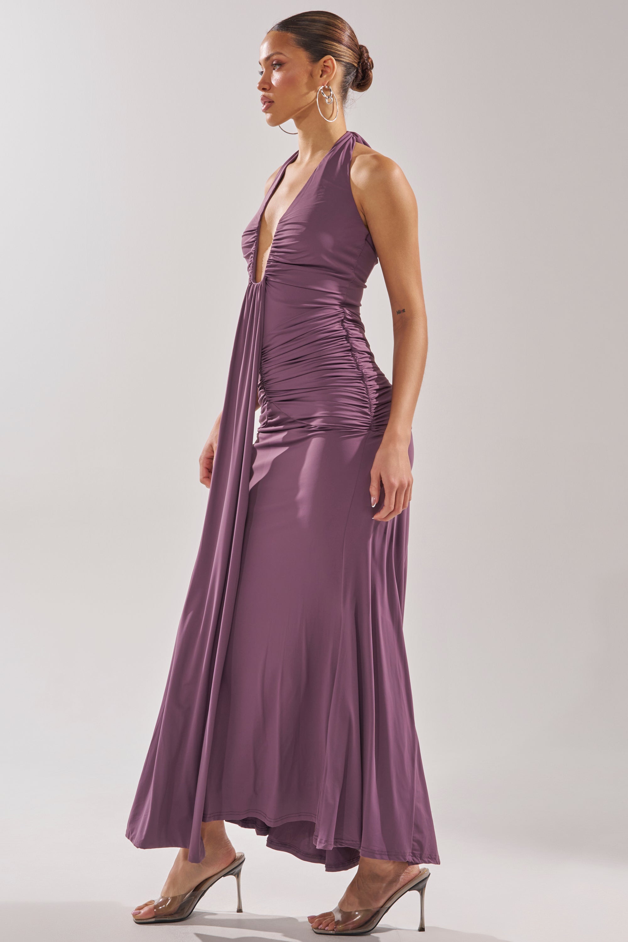 A woman models the ISLAND LOVE MAXI DRESS—a sleeveless, mauve halter maxi with a ruched bodice and deep V-neckline—paired with clear heels as she poses in profile against a plain background. 3