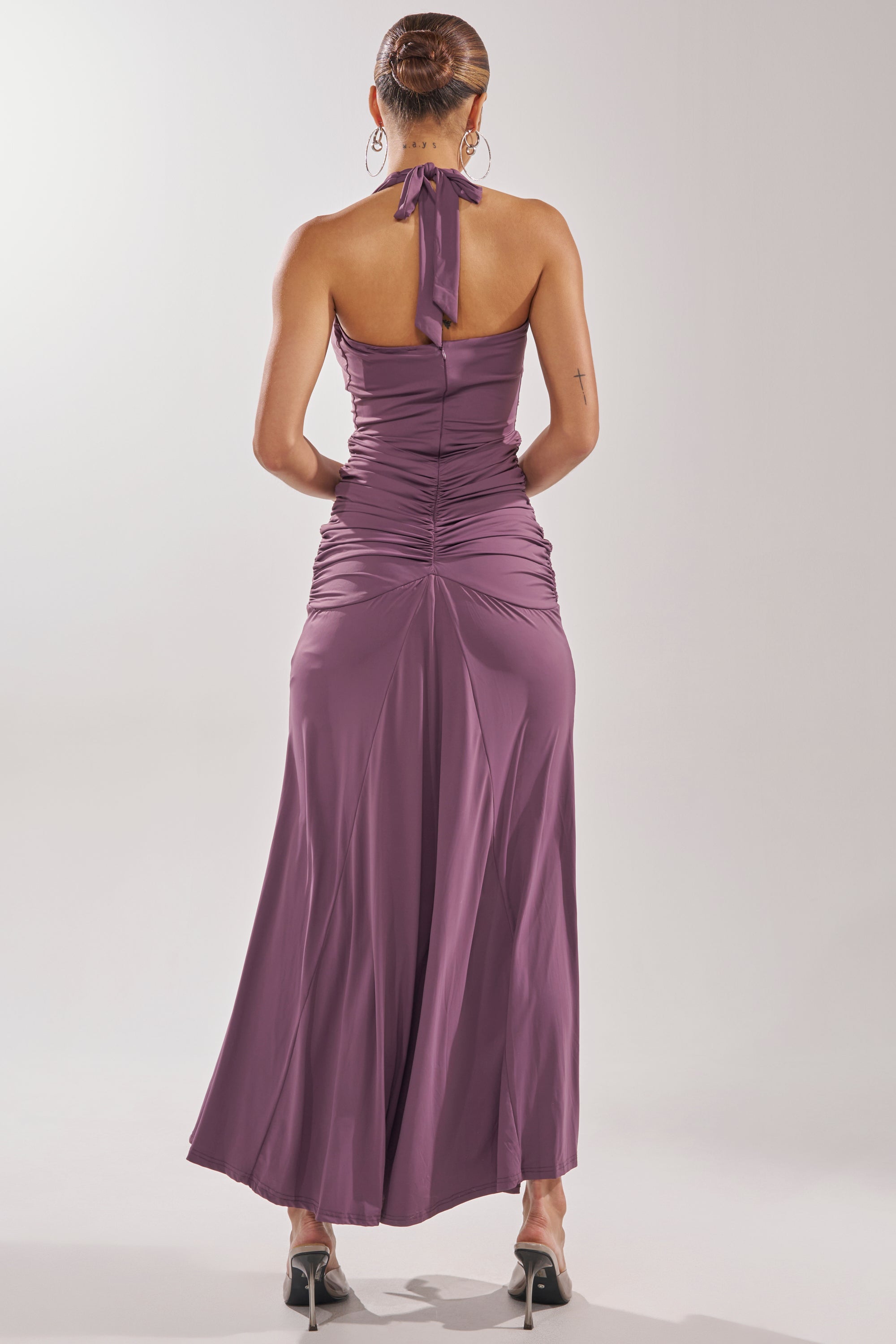 A woman stands with her back turned, wearing the mauve ISLAND LOVE MAXI DRESS featuring ruching and a flowy skirt. Her hair is in a bun, paired with black heels and hoop earrings for a chic vacation look against a light background. 4