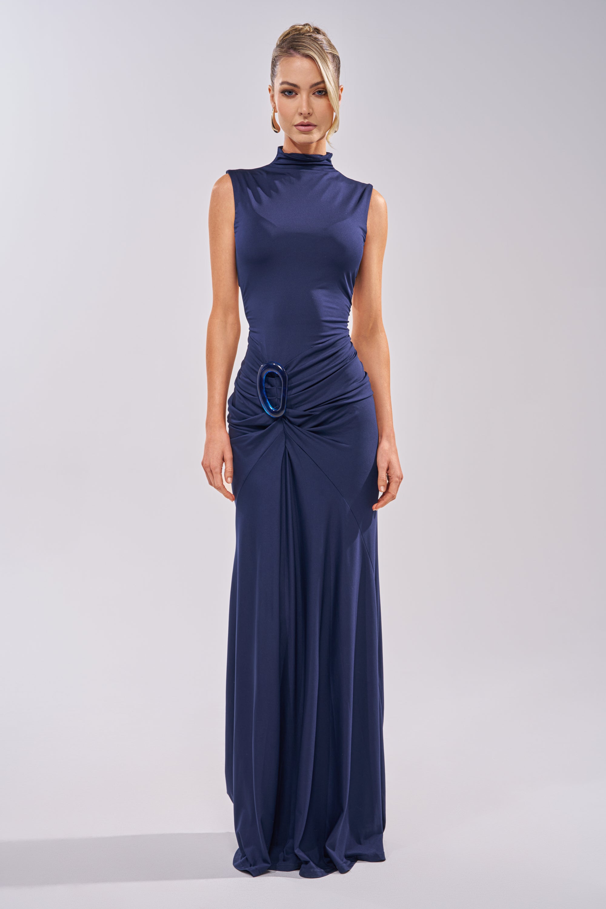 A woman stands against a plain background wearing the BE MY DATE BODYCON MAXI DRESS IN NAVY—sleeveless, floor-length, with a high neckline, gathered waist detail, and accented by a large circular buckle. 1