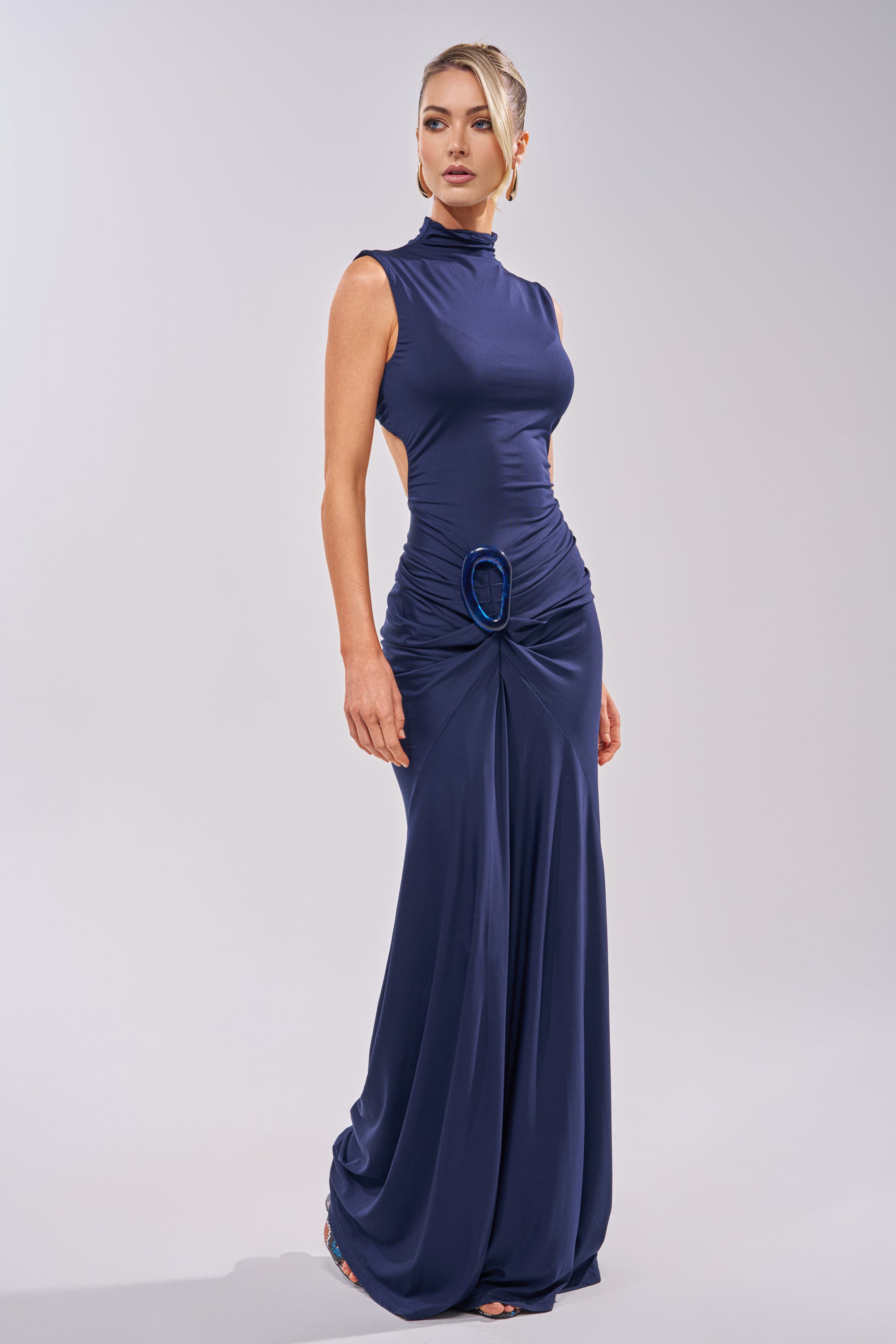 A woman models the BE MY DATE BODYCON MAXI DRESS IN NAVY, a sleeveless, floor-length bodycon dress featuring side cutouts and a gathered waist, posing against a plain light background. 2