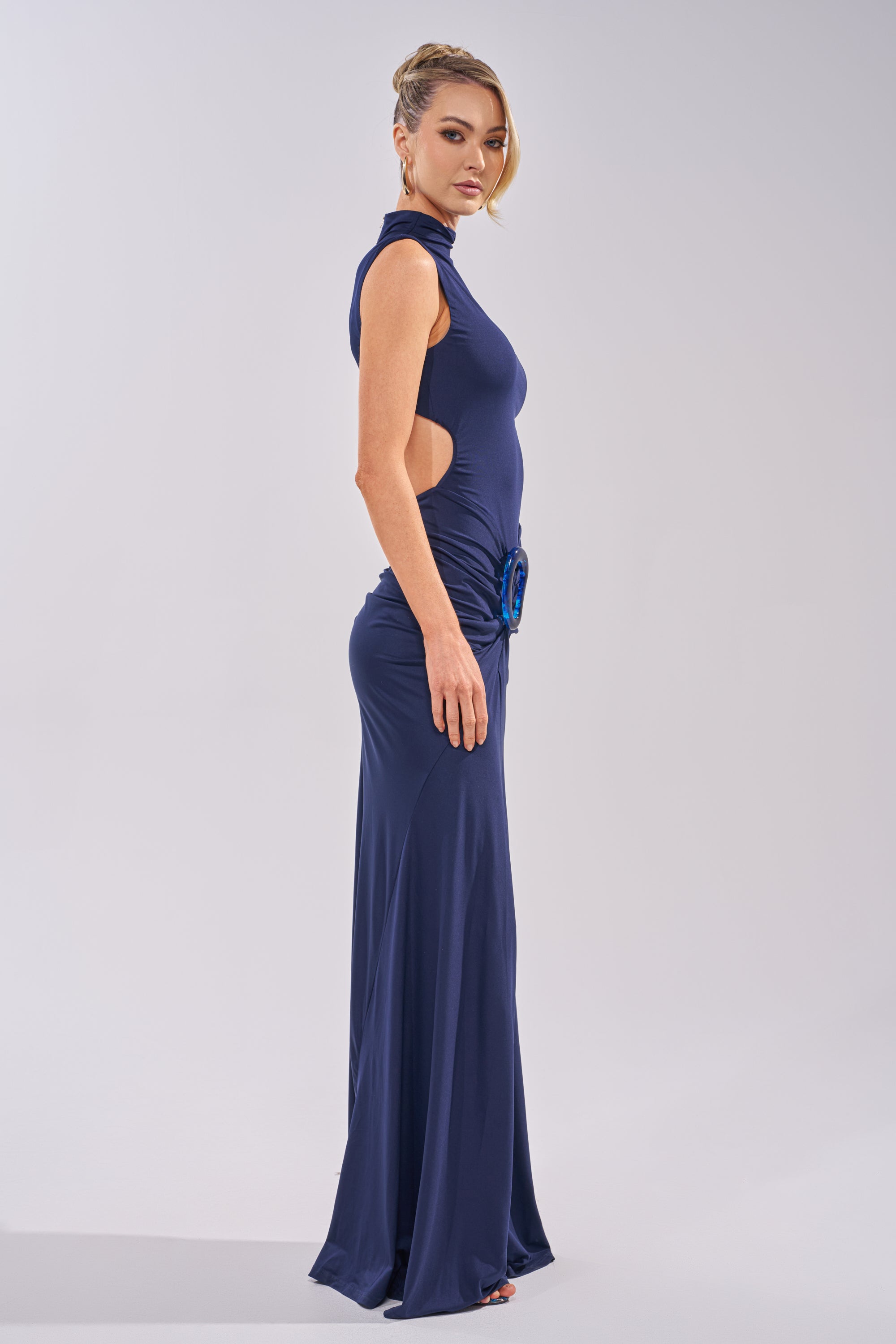 A woman stands sideways against a plain, light background wearing the BE MY DATE BODYCON MAXI DRESS IN NAVY—a sleeveless, floor-length bodycon dress with side cutouts and a decorative hip detail. 3