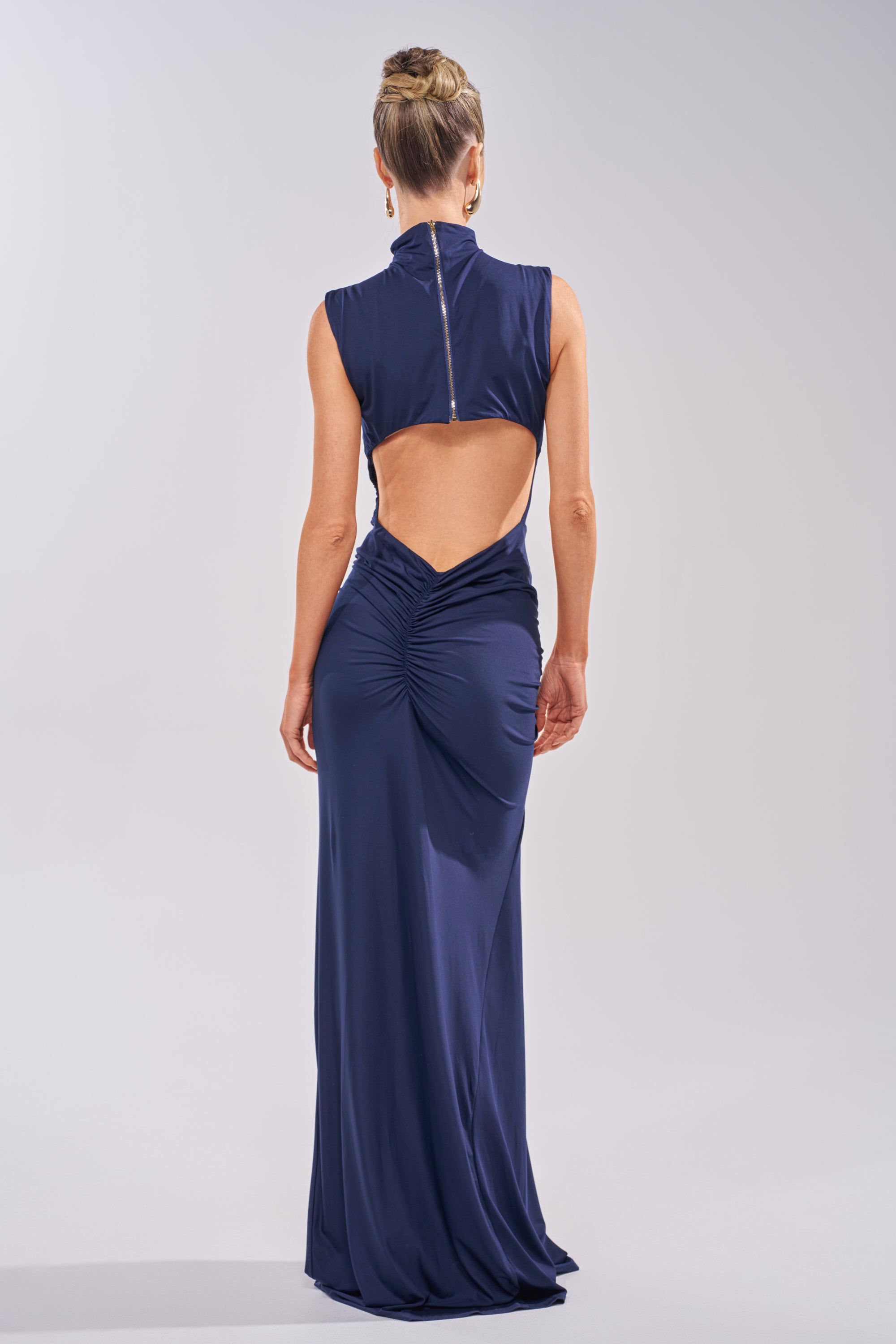 A woman with a neat updo faces away, wearing the BE MY DATE BODYCON MAXI DRESS IN NAVY—a floor-length, sleeveless gown with an open back and ruched waist. The background is light gray. 4