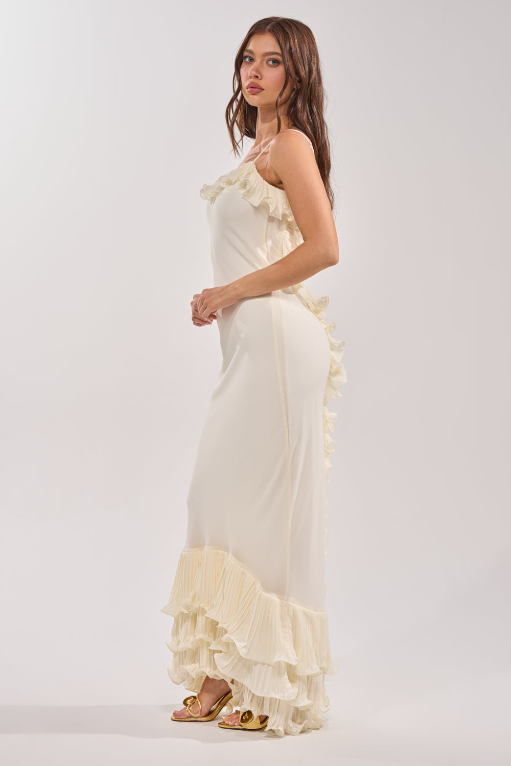 A woman with long brown hair stands sideways in the LOVE ME OR LOSE ME RUFFLE MAXI DRESS IN IVORY, featuring ruffled trim on the hem and side. She pairs the pleated dress with open-toe sandals, gazing just past the camera against a plain background. 2