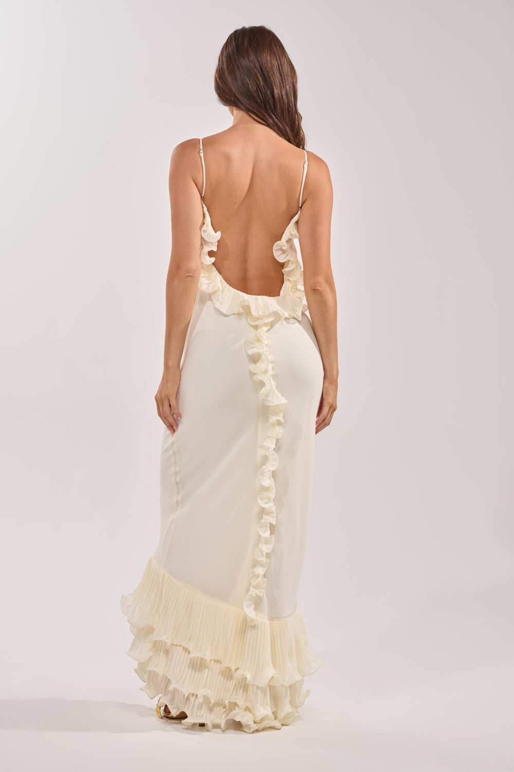 A woman with long brown hair is seen from behind wearing the LOVE ME OR LOSE ME RUFFLE MAXI DRESS IN IVORY, a backless, flowing pleated ruffle dress, set against a plain light background. 3
