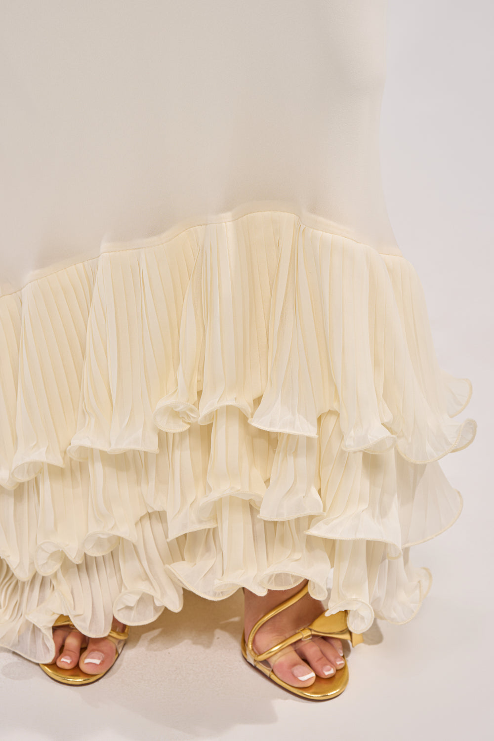 A close-up of the cream pleated ruffle hem on the LOVE ME OR LOSE ME RUFFLE MAXI DRESS IN IVORY, with gold strappy sandals peeking out beneath. 5