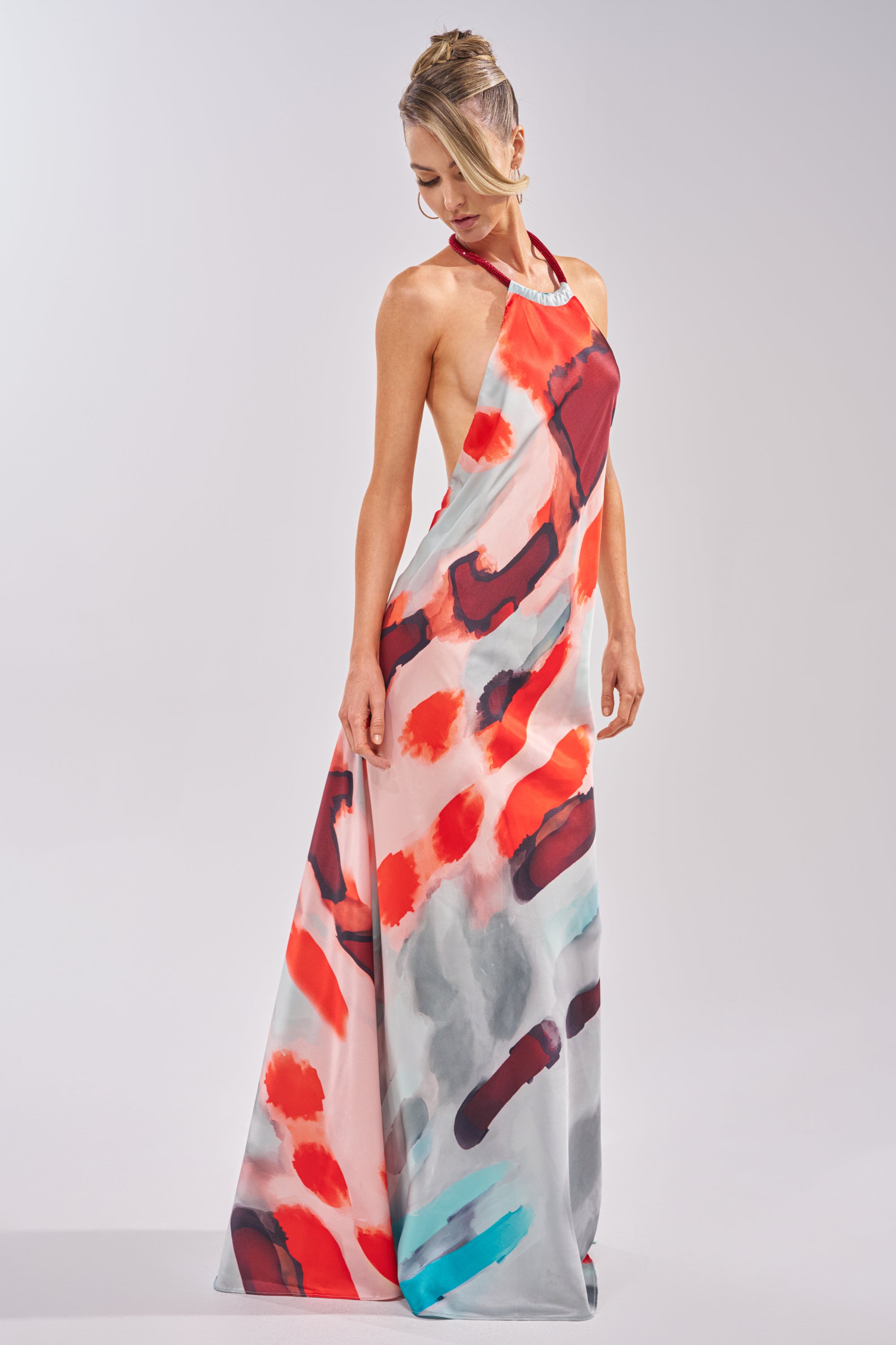 A woman wearing the CHIC IS ME SATIN MAXI DRESS IN MULTI, featuring a halter neckline and an abstract red, orange, black, and blue print, stands against a plain light background looking down. 0