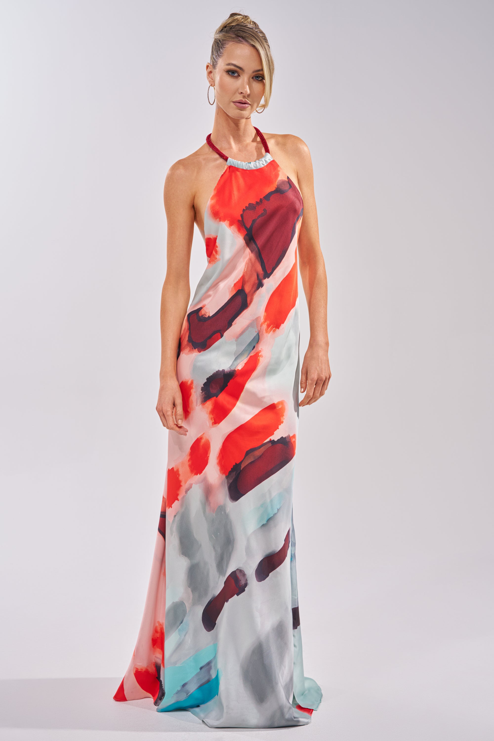 A woman wears the CHIC IS ME SATIN MAXI DRESS IN MULTI, a flowing halter dress with bold red, black, turquoise, and gray abstract patterns, standing against a plain light background. 1