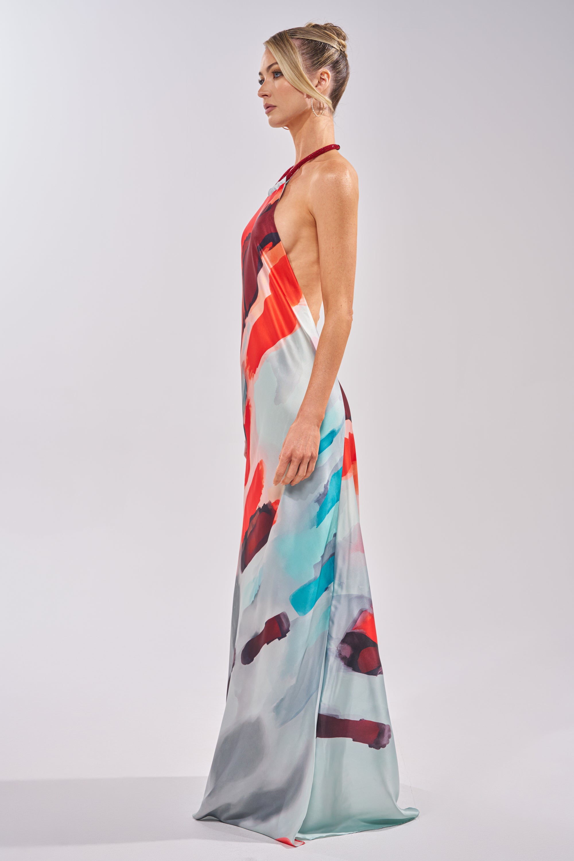 A woman in a sleek bun poses in profile, wearing the CHIC IS ME SATIN MAXI DRESS IN MULTI—an abstract print halter dress with a flowing, floor-length silhouette in red, blue, and gray tones against a plain light background. 3