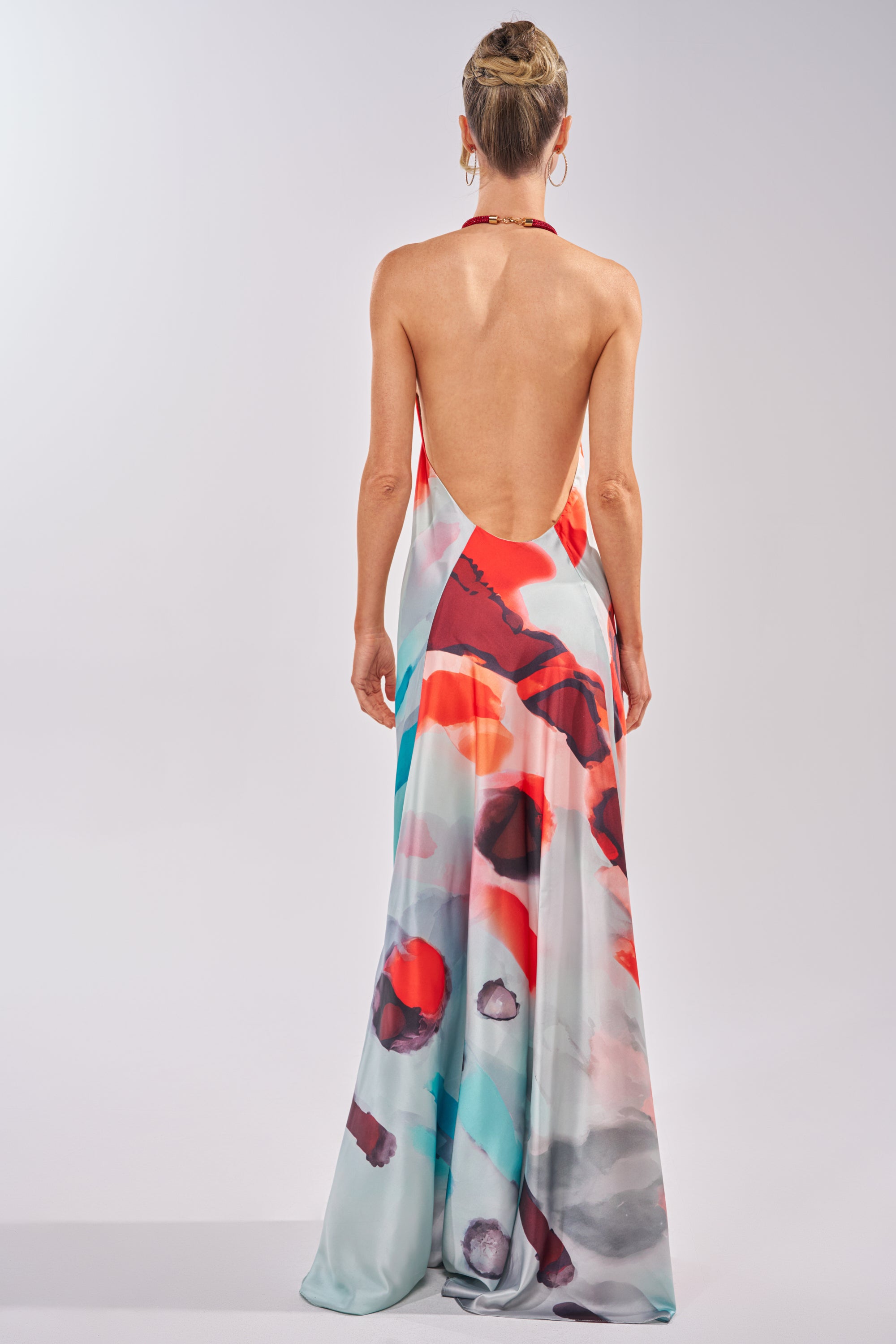 A woman with her hair in a neat bun faces away, wearing the CHIC IS ME Satin Maxi Dress in Multi. This abstract print halter dress features red, blue, gray, and white hues, a flowing silhouette, open back, and an eye-catching satin finish. 4