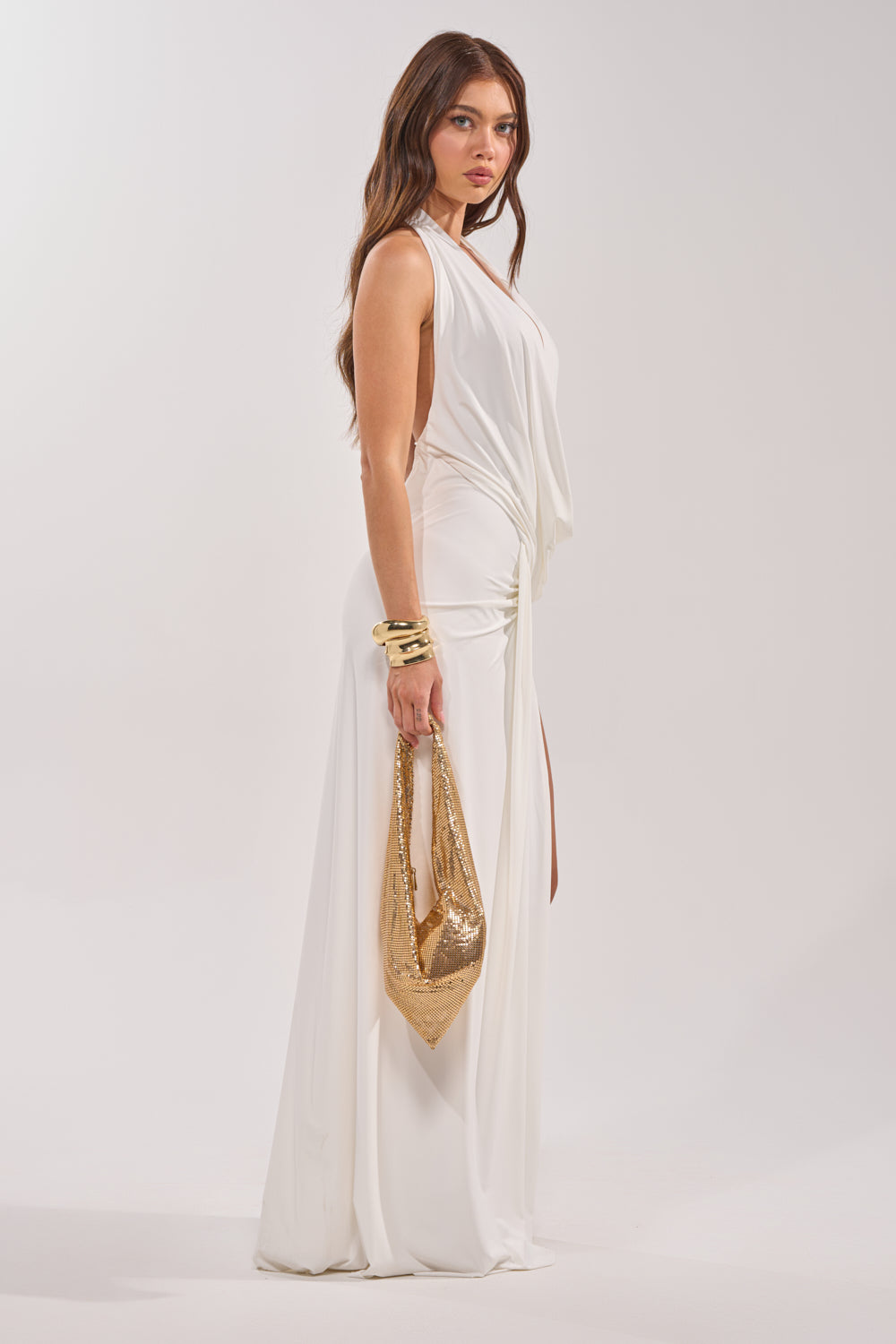 A woman wears the HEAD TURNING HALTER MAXI DRESS IN WHITE, holding a gold handbag and sporting gold bangles. She poses against a plain light background, glancing slightly over her shoulder. 5