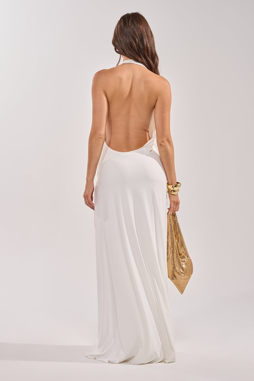 A woman with long brown hair faces away, showcasing the HEAD TURNING HALTER MAXI DRESS IN WHITE—ideal for formal occasions. She holds a gold sequined handbag and wears chunky gold bracelets against a plain, light background. 6