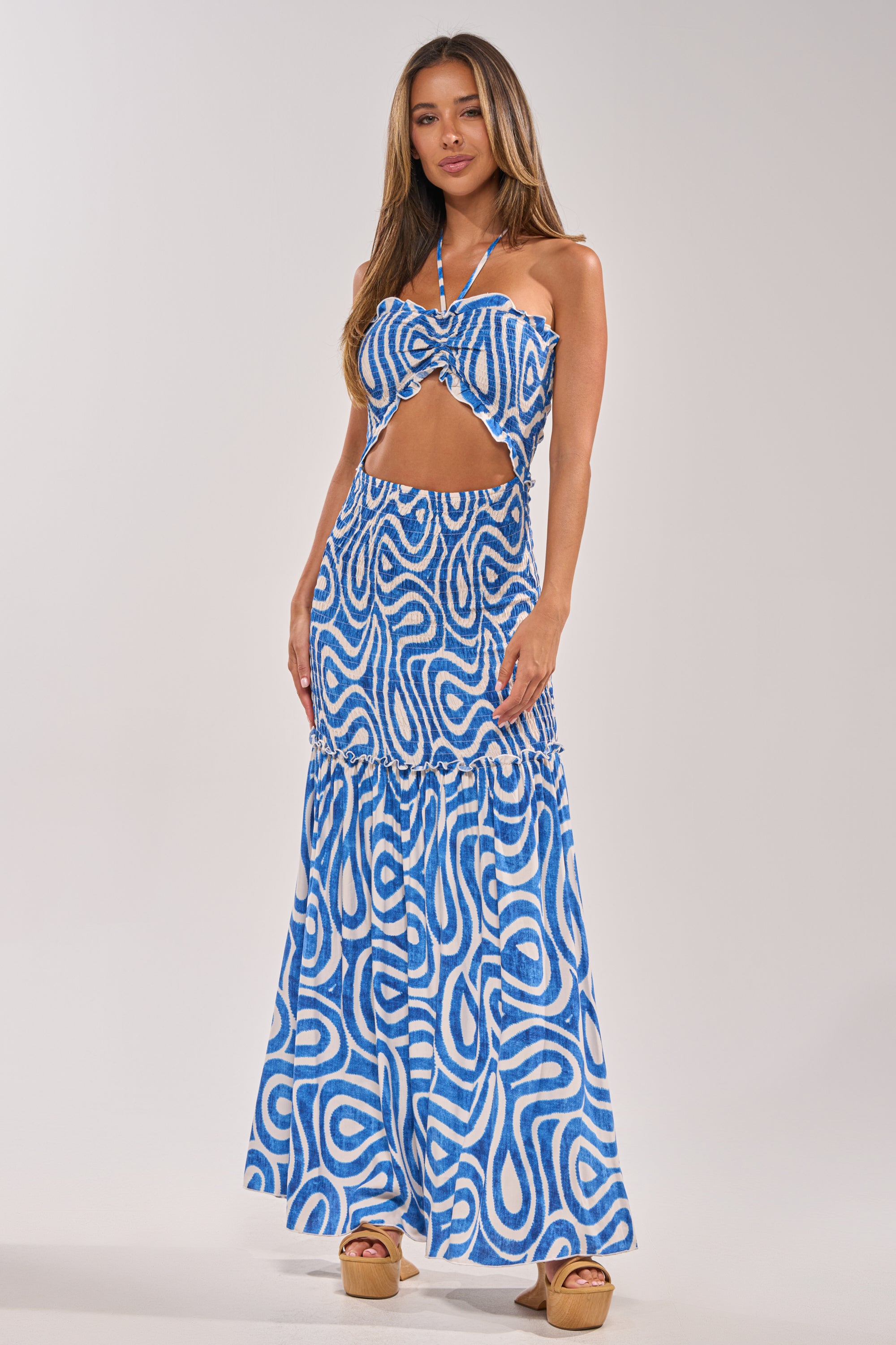 A woman models the SUGAR AND SPICE PRINTED MAXI DRESS IN BLUE MULTI, featuring a front cutout and halter neckline, paired with tan platform sandals—an ideal choice for summer. 1