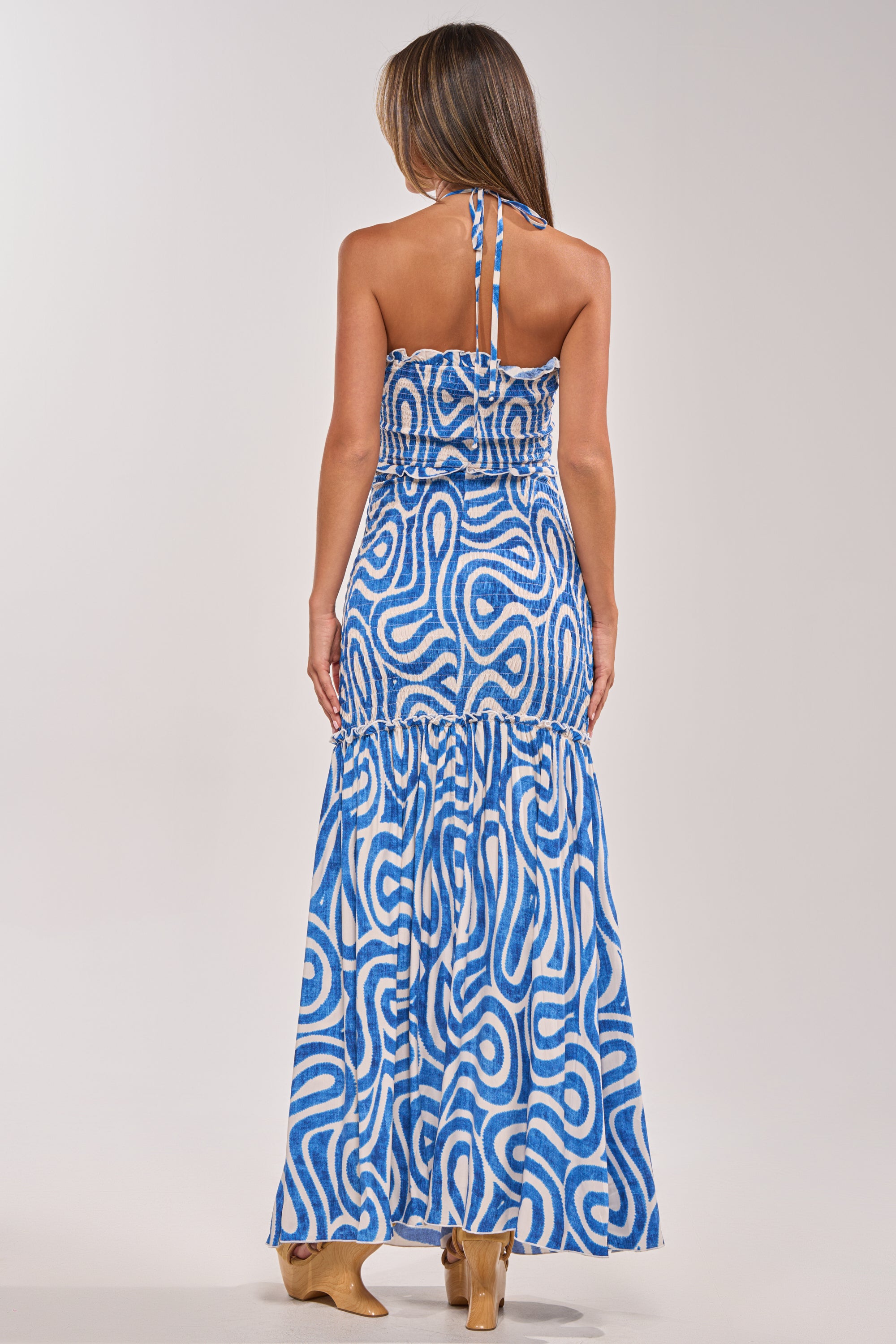 A woman with long brown hair faces away, modeling the SUGAR AND SPICE PRINTED MAXI DRESS IN BLUE MULTI—featuring an abstract swirl pattern and flared hem—paired with tan heeled boots against a plain light background. 3