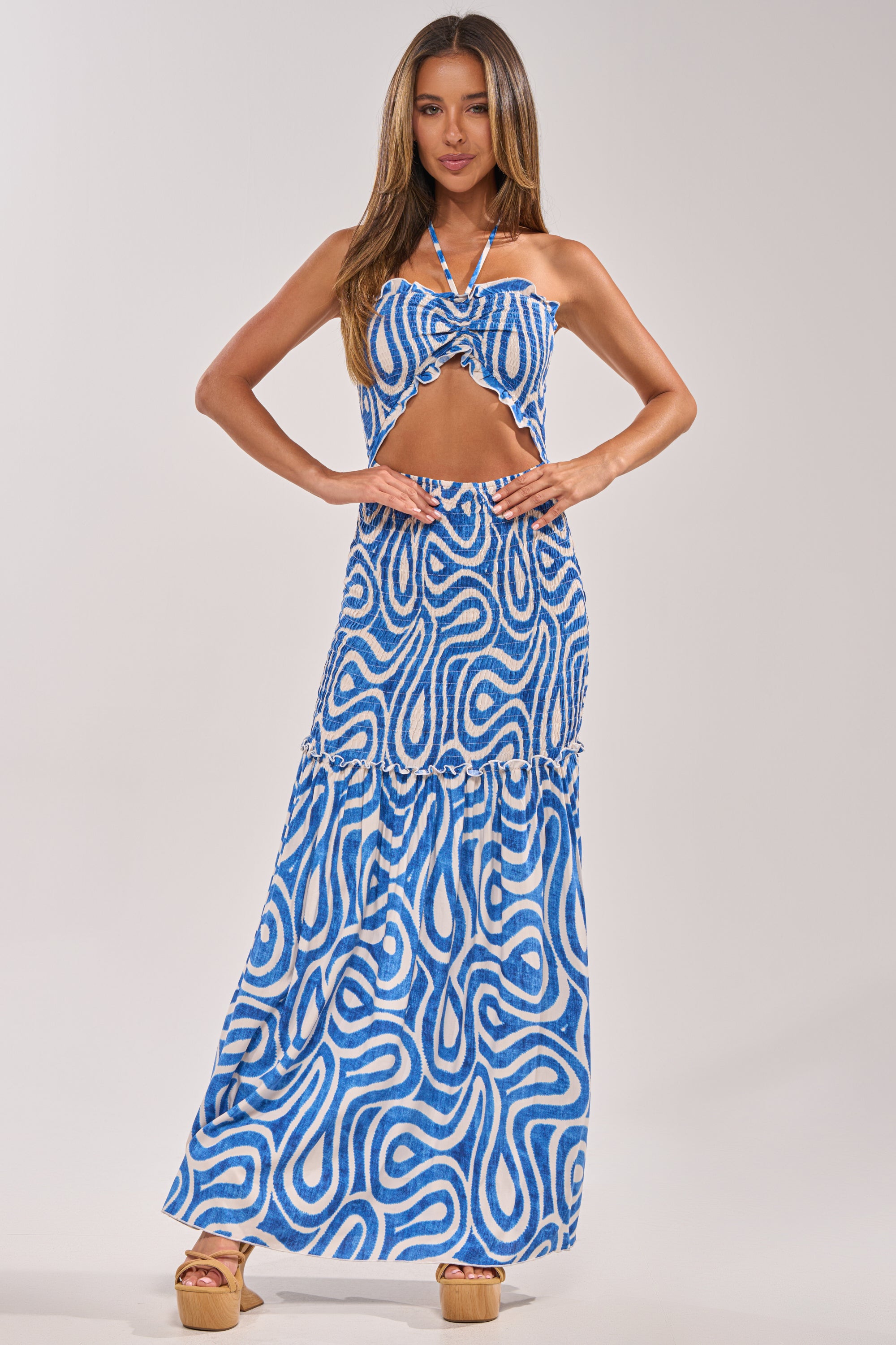 A woman models the SUGAR AND SPICE PRINTED MAXI DRESS IN BLUE MULTI, featuring a halter neckline, abstract print, front cut-out, and tiered skirt. She pairs it with light brown platform sandals against a plain background. 4