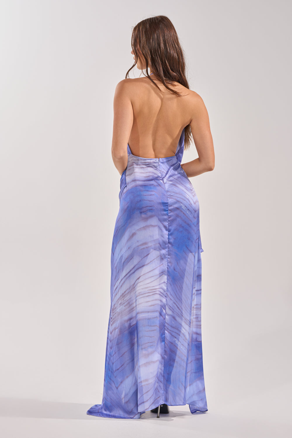 A woman with long brown hair models the PLAY NICE HIGH-LOW DRESS IN PURPLE MULTI, featuring a backless, floor-length design and vibrant patterns—ideal for summer or beach styling against a light background. 4