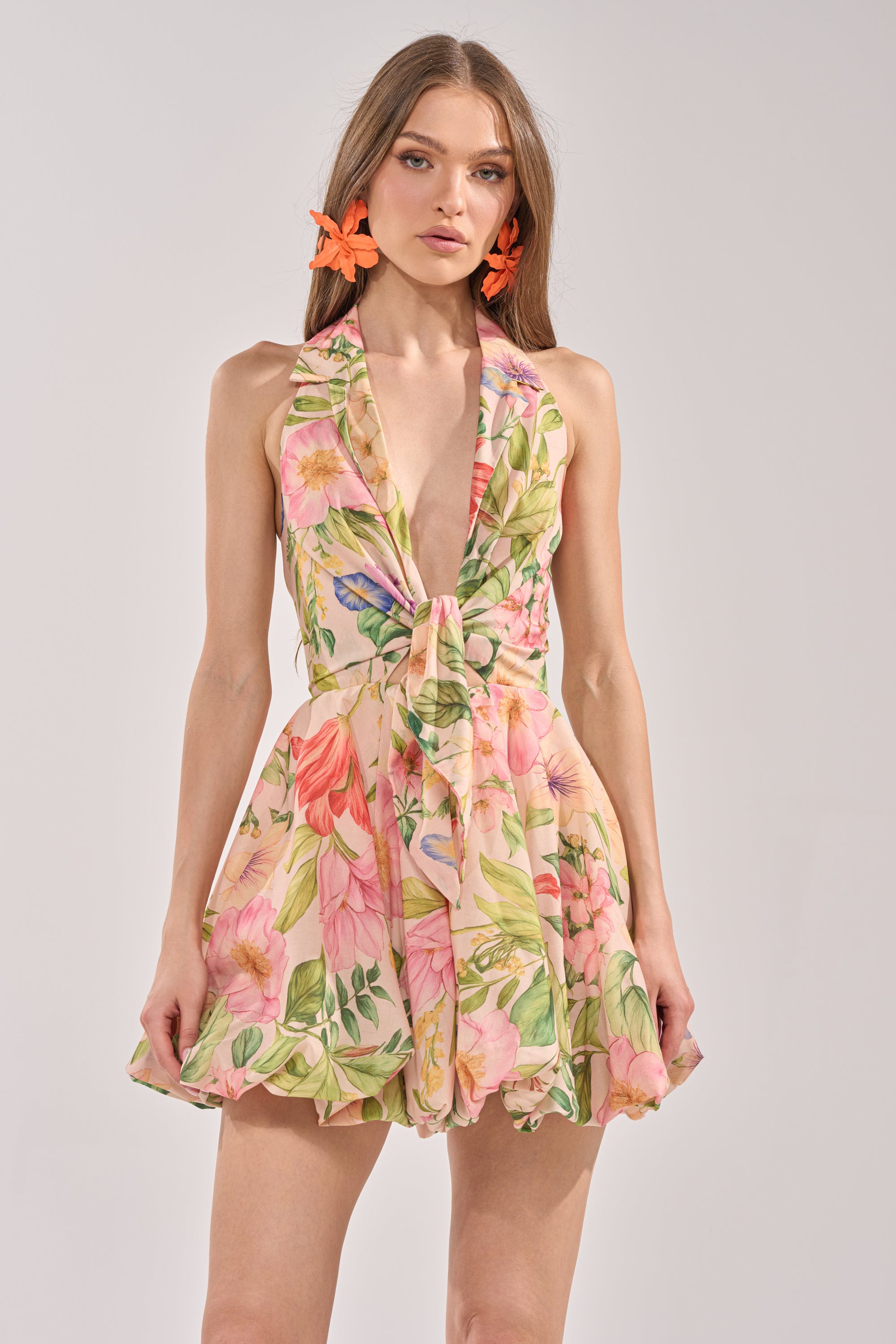 A woman stands against a plain light background in the SOFT LAUNCH FLORAL MINI DRESS IN BEIGE, featuring a sleeveless design, deep V-neckline, and flared skirt. She accessorizes with orange flower earrings for a chic vacation look.