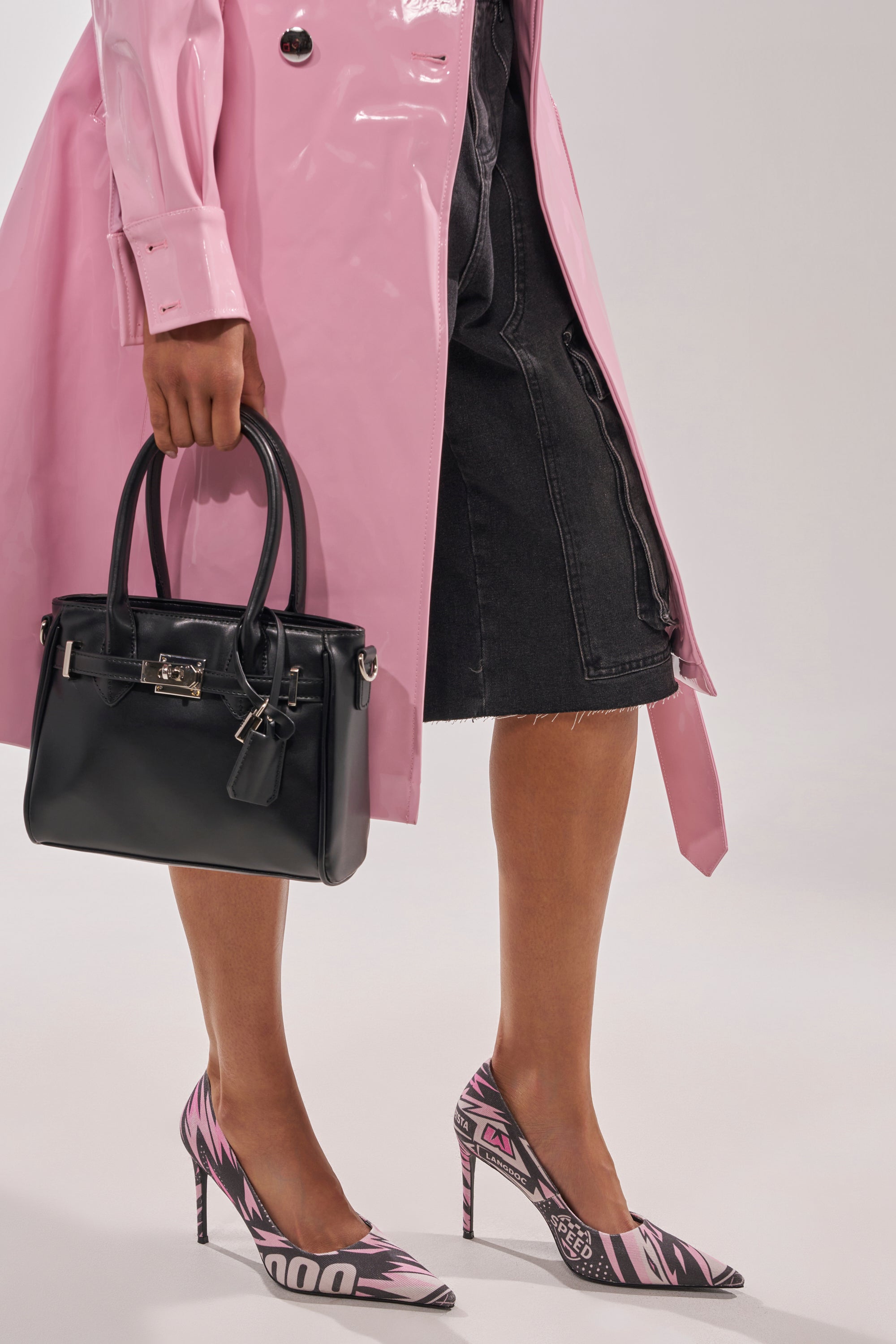 A person in a shiny pink trench coat and black knee-length denim shorts holds a black handbag and wears AZALEA WANG SPORT PRINTED STILETTO POINTED TOE PUMPS. The image is cropped at the upper thigh. 0