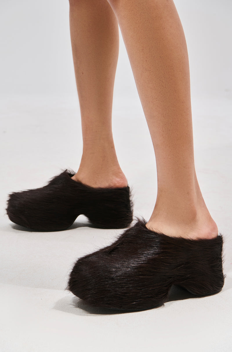 A person stands on a light floor with bare legs visible, wearing the AZALEA WANG LEESHA CHOCOLATE PONY HAIR CLOG slippers against a plain background.