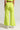 A person is wearing the SWEET AND SPICY WIDE LEG PANT in lime green, which is high-rise and semi-stretchy, accompanied by a matching top and silver high heels. The image captures the person from behind against a plain, light-colored background.