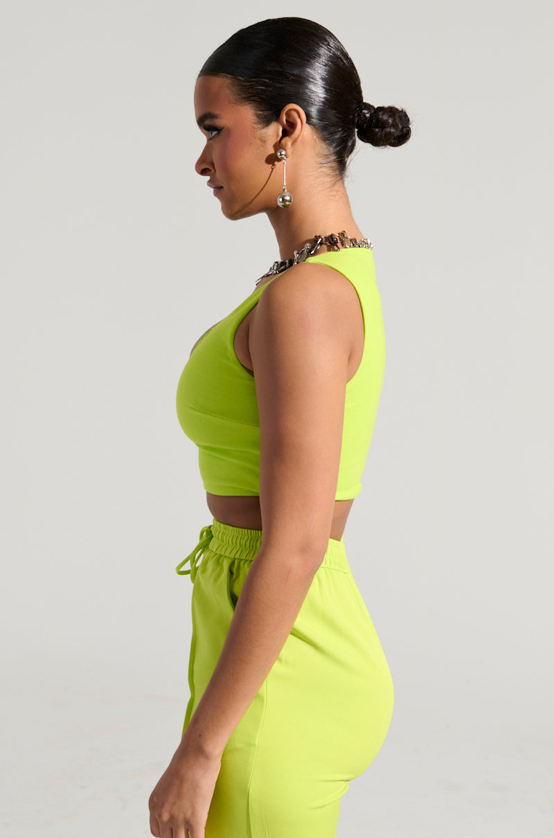 In profile, a person with their hair in a bun showcases the SWEET AND SPICY TANK—an eye-catching bright green sleeveless crop top featuring a plunging v-neckline, paired with matching pants. The look is completed with hoop earrings and a chunky chain necklace against a plain light gray background.