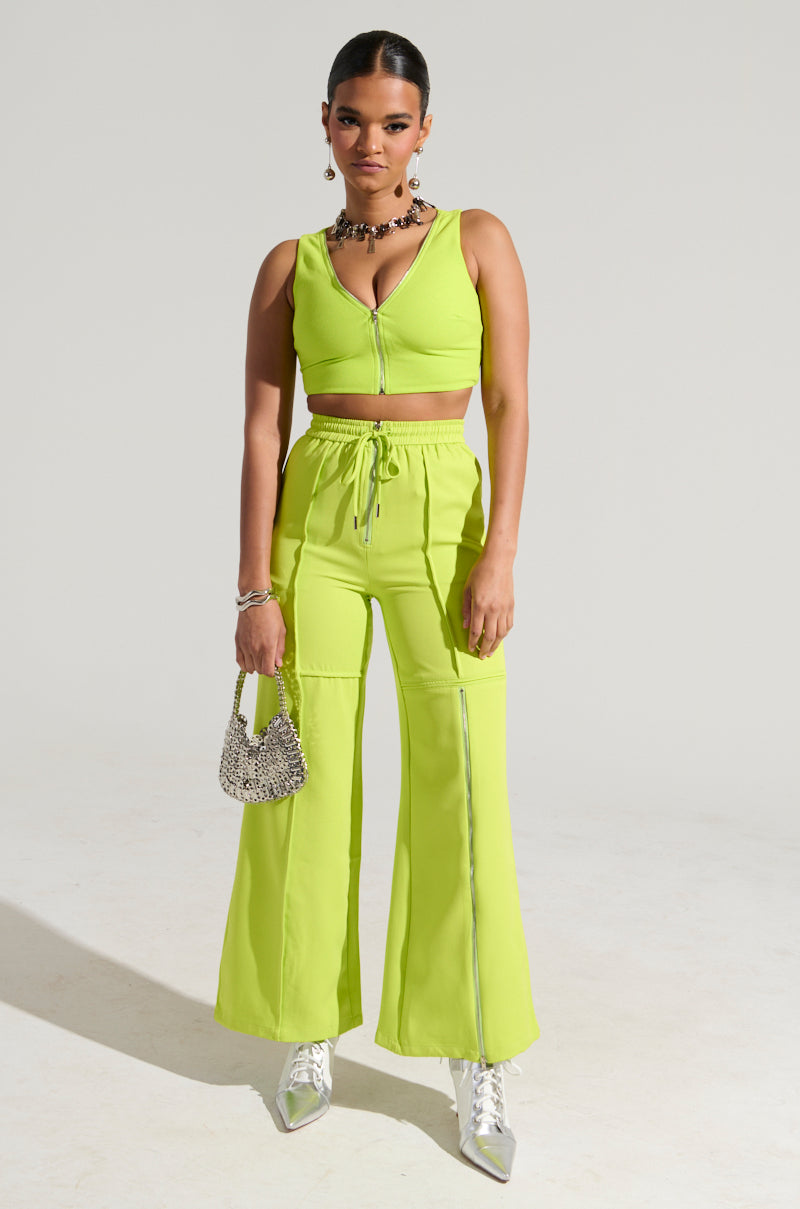 A person poses against a plain backdrop, styled in a vibrant lime green ensemble featuring the SWEET AND SPICY TANK and matching cropped wide-legged pants. They accessorize with a small silver beaded handbag and metallic silver ankle boots. Their sleek hairstyle is accented by a necklace.