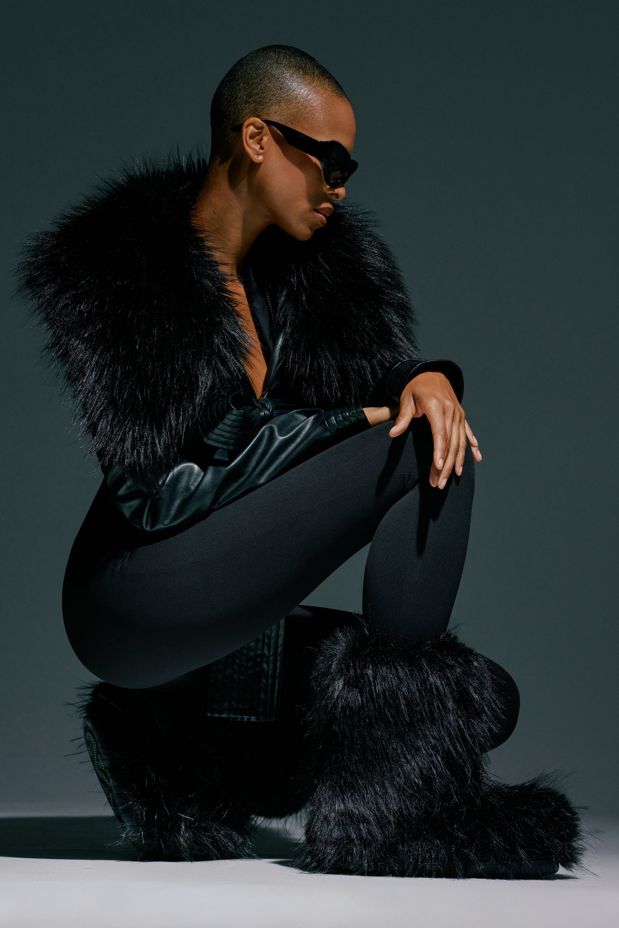 A person wearing dark sunglasses, a black faux fur coat, black leggings, and the AZALEA WANG LENIX BLACK FLAT FUR BOOTIE crouches in a stylish pose against a plain dark background.