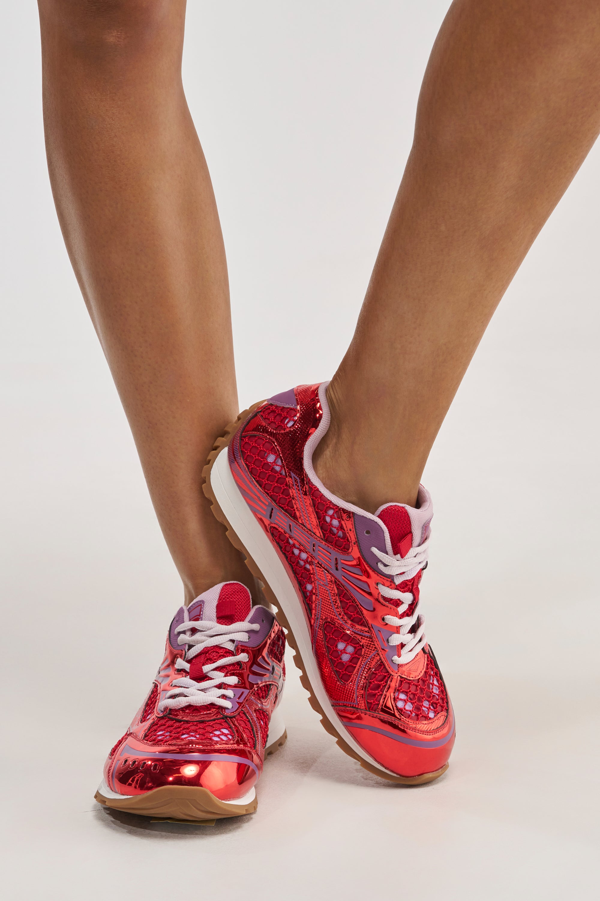 A person wearing the AKIRA GOLD LENIYA RED SNEAKER—with a metallic red sporty-chic design, white laces, and brown soles—stands with legs crossed on a plain white background.