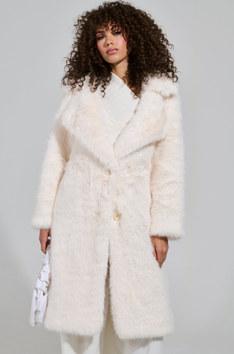 A person with curly hair is wearing the LENOX FAUX FUR TRENCH IN IVORY over a white outfit. They are holding a white handbag, and the background is plain and light-colored.