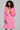 A woman with curly hair wears the LENOX FAUX FUR TRENCH IN PINK over a lime green dress. She holds a beaded clutch and accessorizes with layered necklaces, perfectly complementing her high-quality faux fur ensemble. She stands strikingly against a plain backdrop.